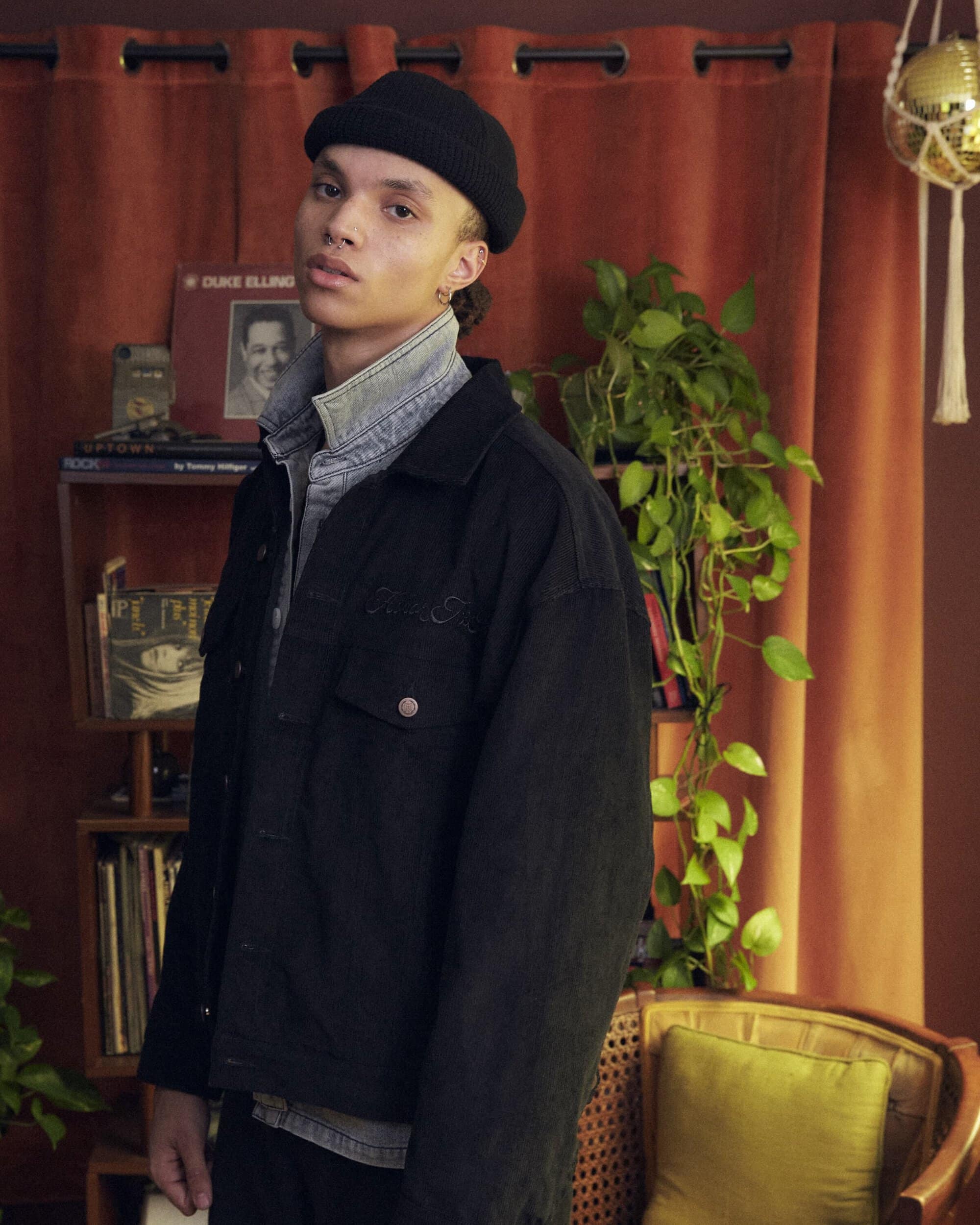 Young adult man wearing a black beanie and oversized jacket poses in a room with vintage decor and warm, inviting tones.