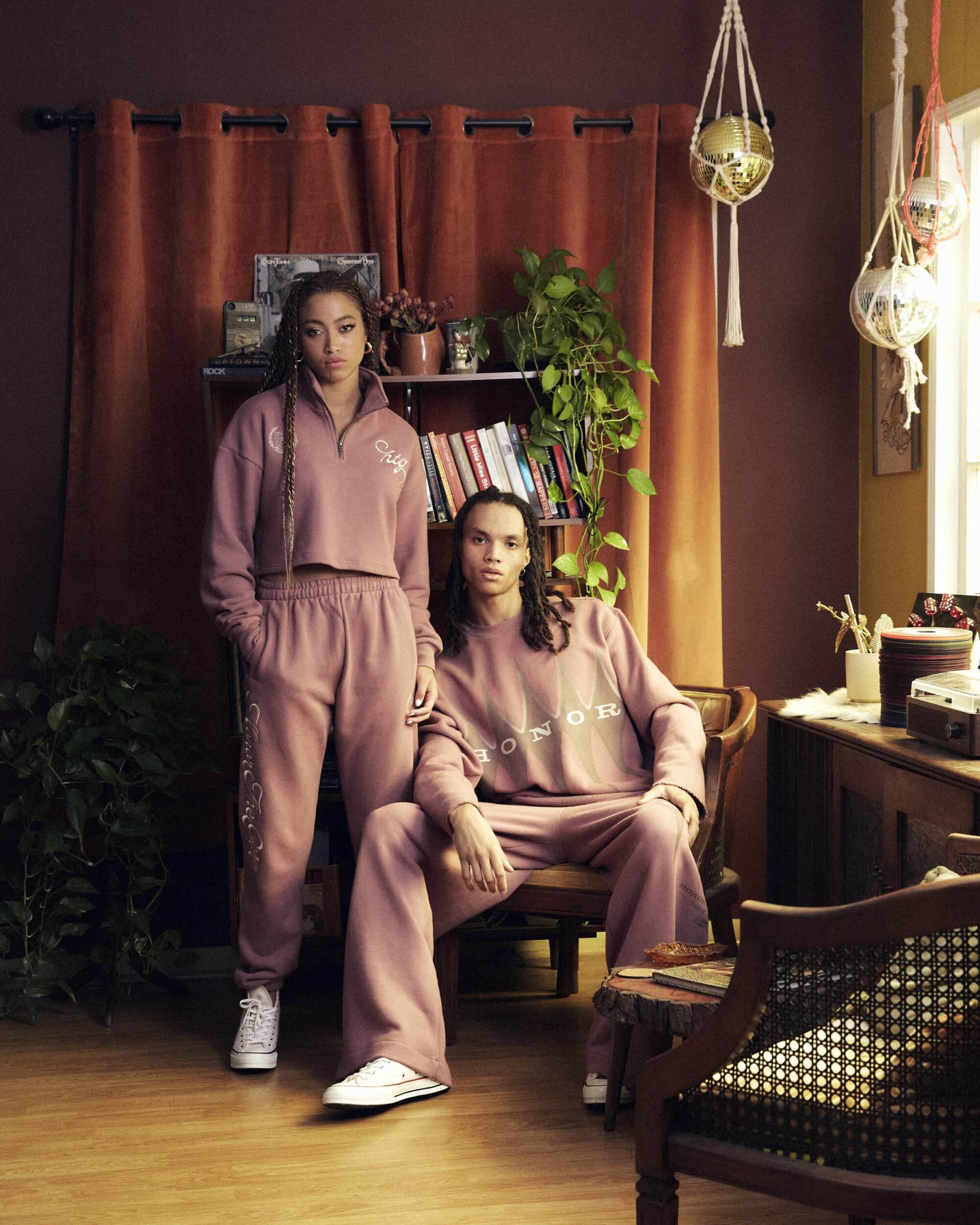 Two models wearing matching dusty rose sweatsuits with white sneakers in a styled interior with a bookcase and plants.
