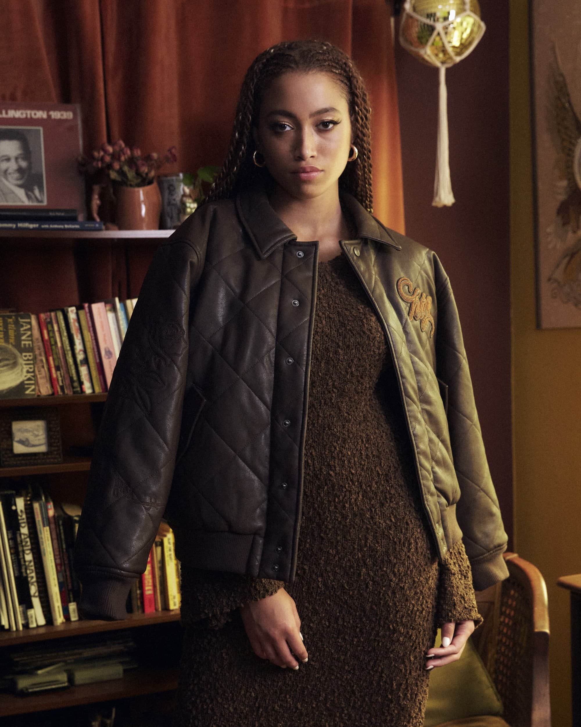 Woman in brown leather jacket and knitted dress poses indoors near a bookshelf with warm, natural lighting and a nostalgic aesthetic.