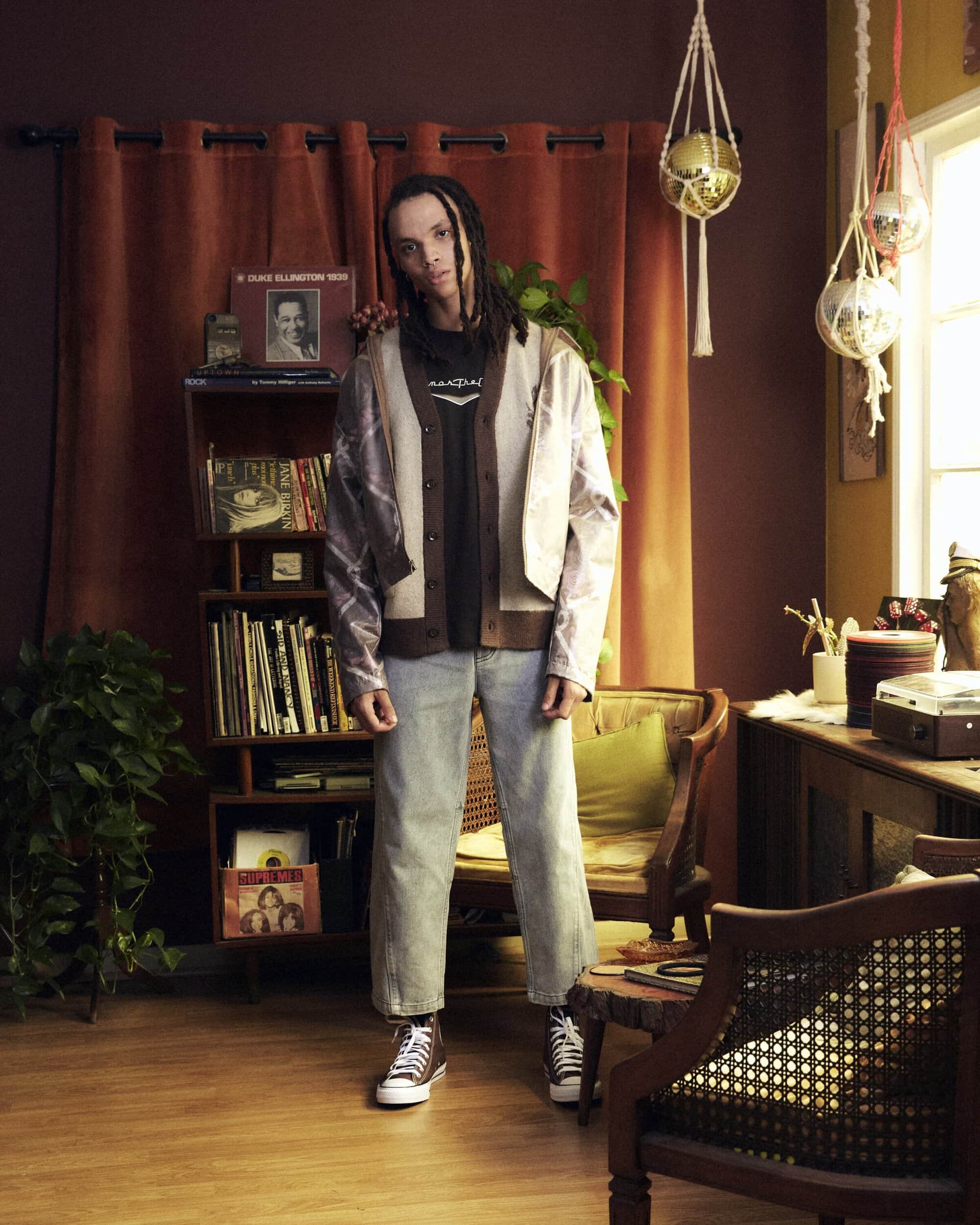 Full shot of a person with dreadlocks, wearing a cardigan and jeans, posing in a room with vintage decor and plants. 