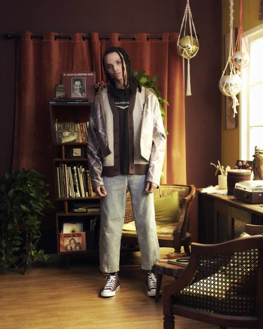 Full shot of a person with dreadlocks, wearing a cardigan and jeans, posing in a room with vintage decor and plants. 