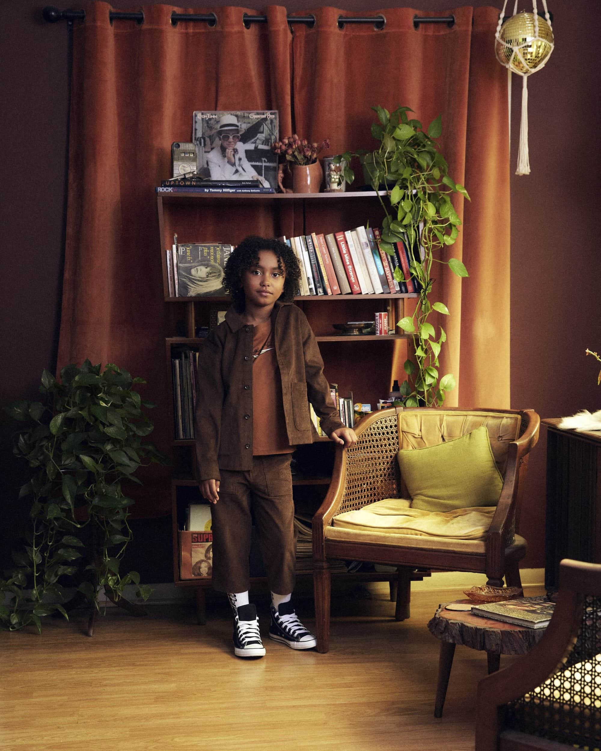 A young child poses in a brown jacket and pants near a bookshelf, rattan chair, and houseplants in a naturally lit, earth-toned room.