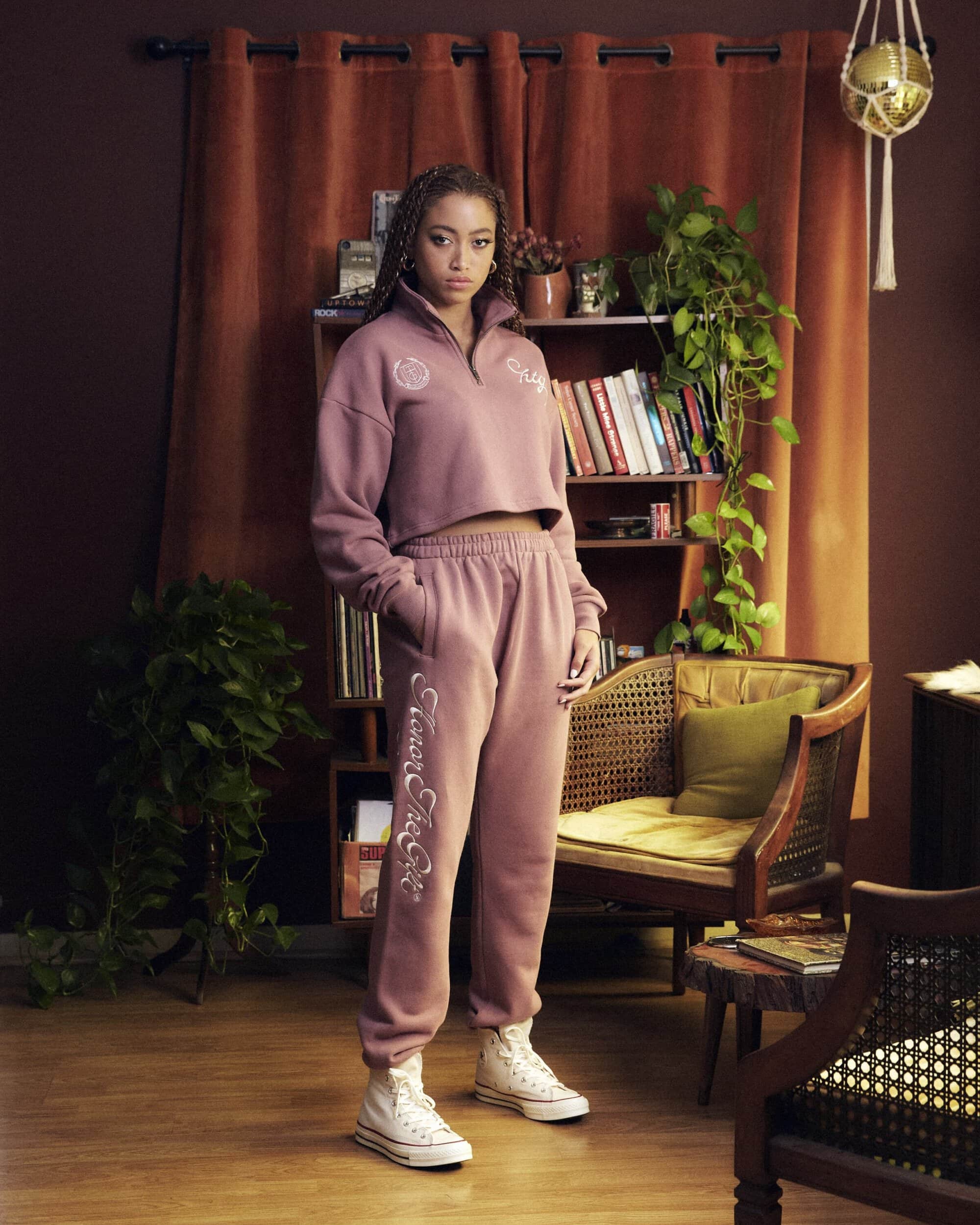 Woman in matching mauve sweat suit poses in front of bookcase with houseplants and dark orange curtain in a softly lit indoor setting.