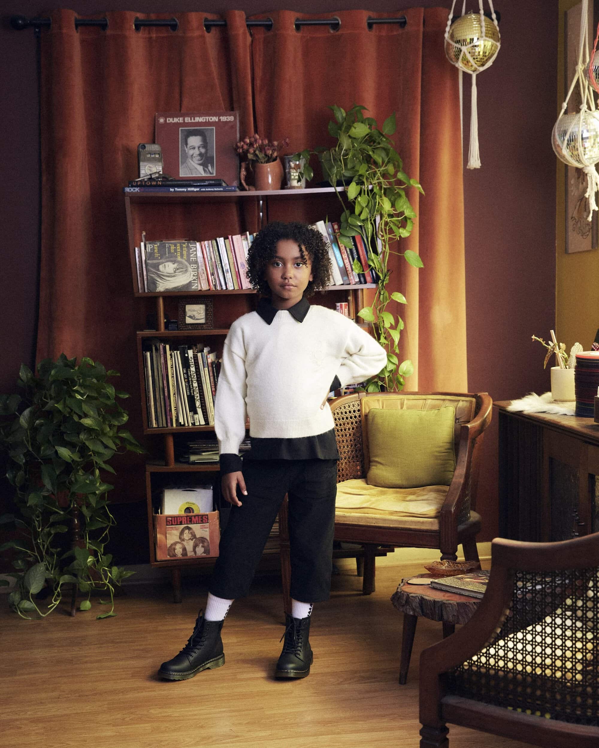 A young person stands in a room with records and plants, wearing a white sweater, black culottes, and black boots, posing confidently.