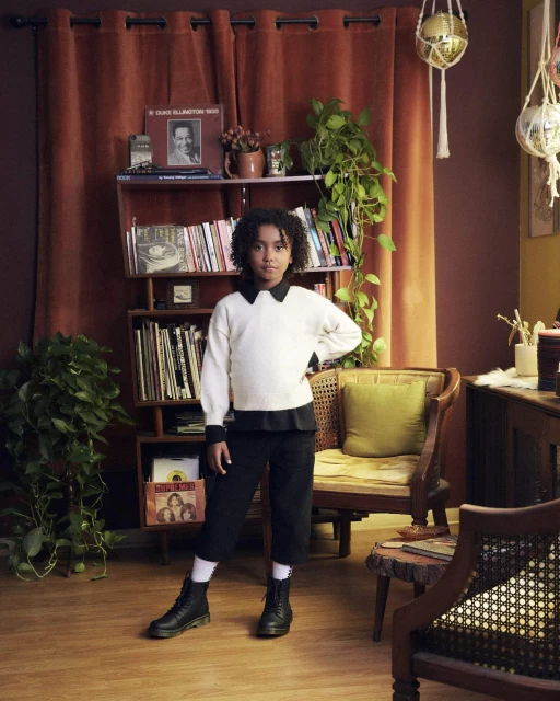 A young person stands in a room with records and plants, wearing a white sweater, black culottes, and black boots, posing confidently.
