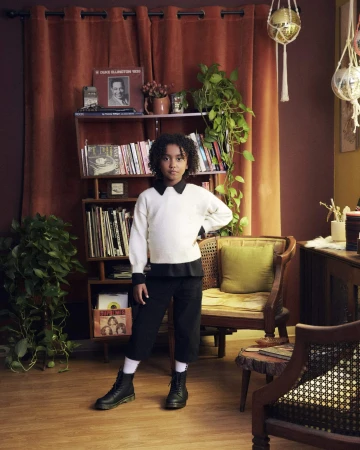 A young person stands in a room with records and plants, wearing a white sweater, black culottes, and black boots, posing confidently.