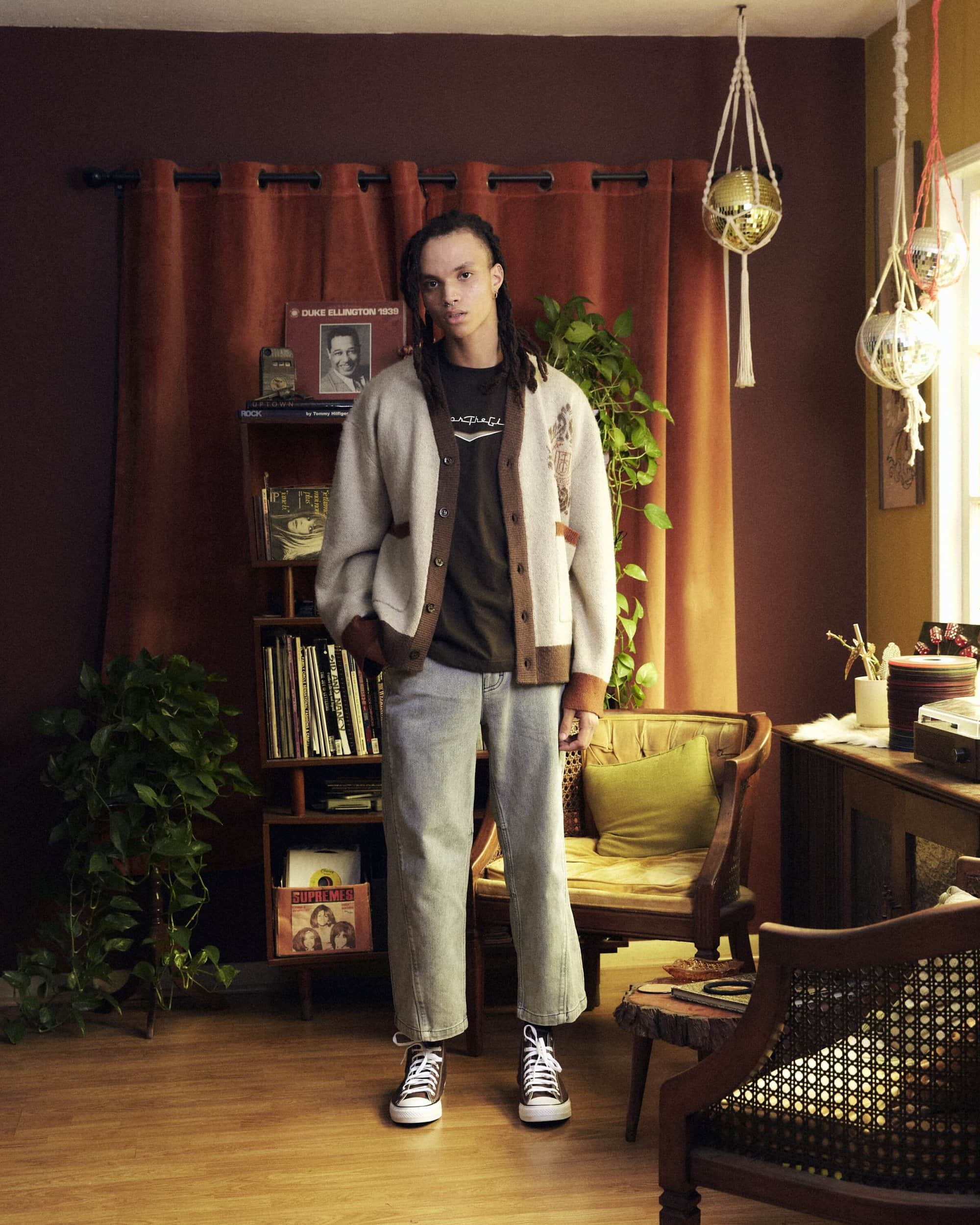 Man stands in a room with a record shelf wearing a cardigan, t-shirt, jeans, and sneakers. Houseplants and vintage furniture visible.