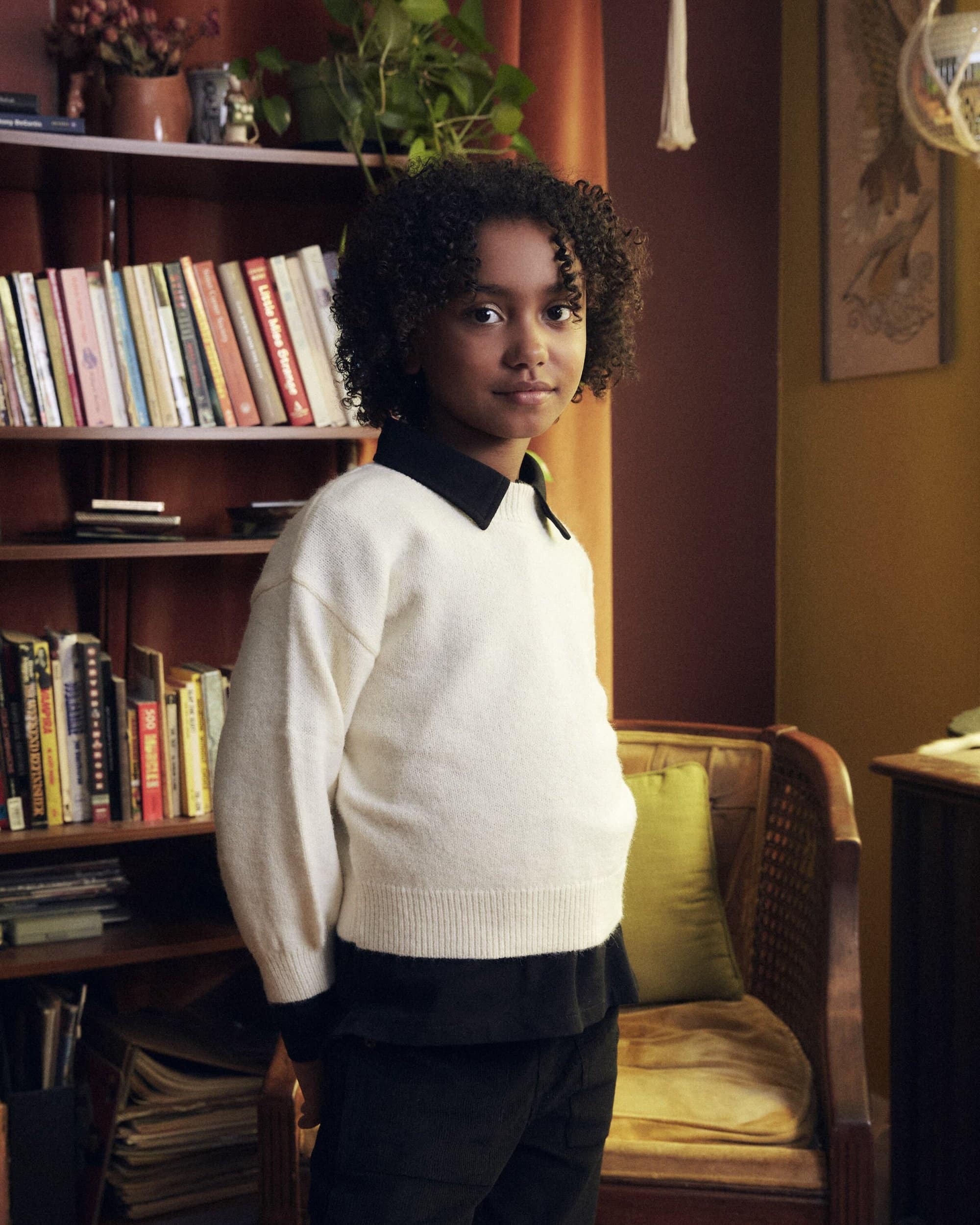 Portrait of a preteen child in a light sweater posing indoors near a bookcase and chair in a cozy, warm-toned domestic setting.