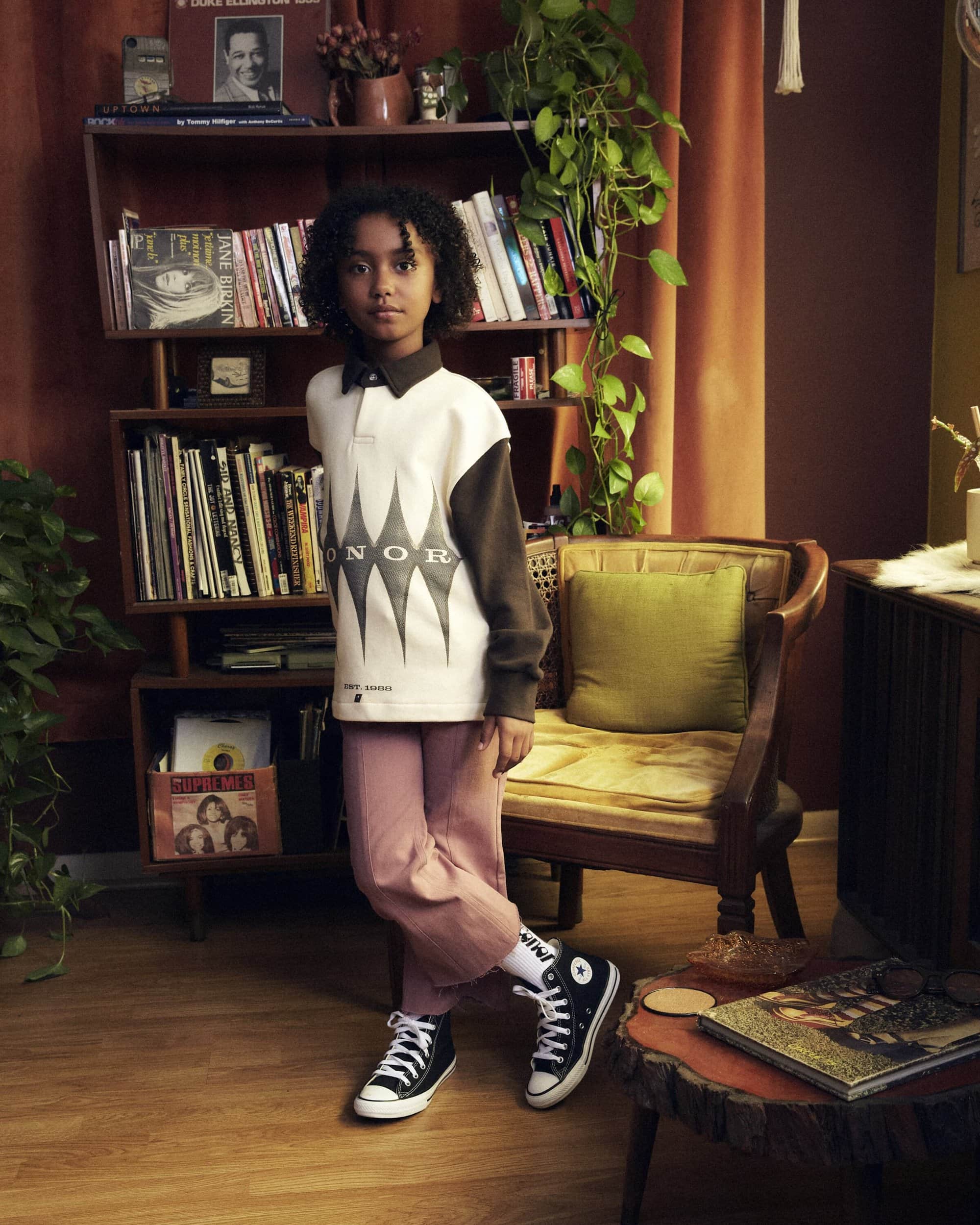 Young child poses in black and white sneakers, light pink pants, and a white shirt, indoors in a room with a bookcase, and chair.