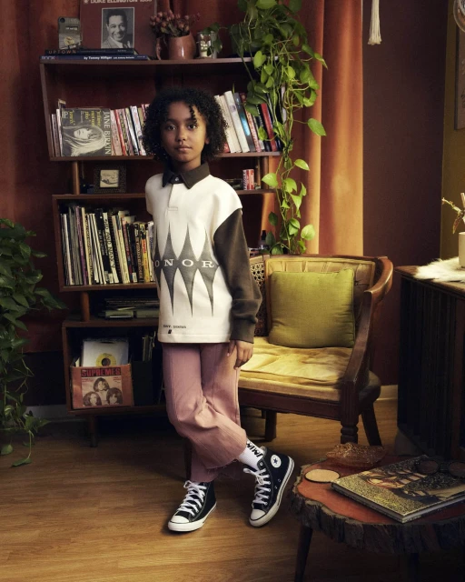 Young child poses in black and white sneakers, light pink pants, and a white shirt, indoors in a room with a bookcase, and chair.