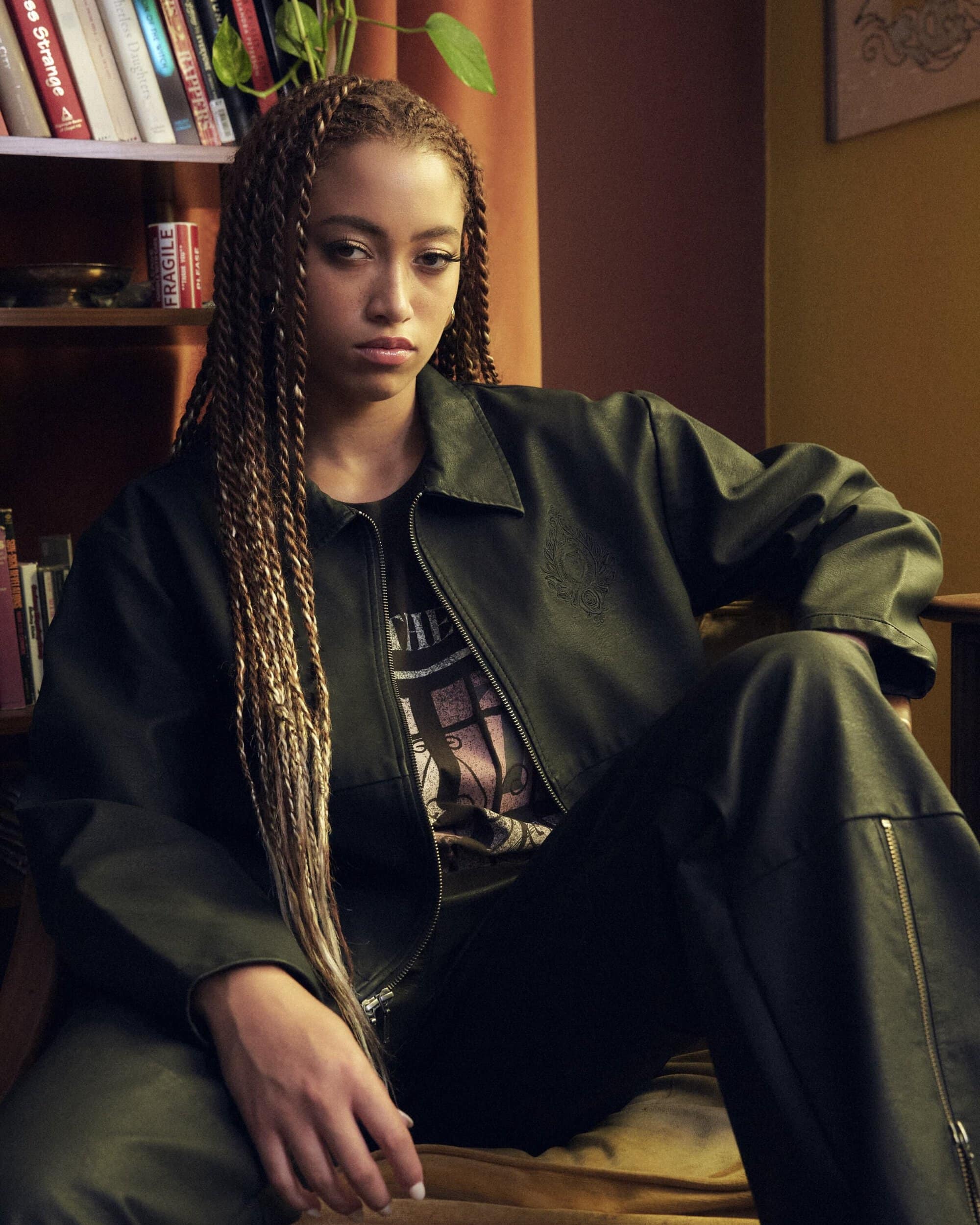 Young woman with long braided hair wearing a dark jacket and trousers sitting indoors by a bookshelf