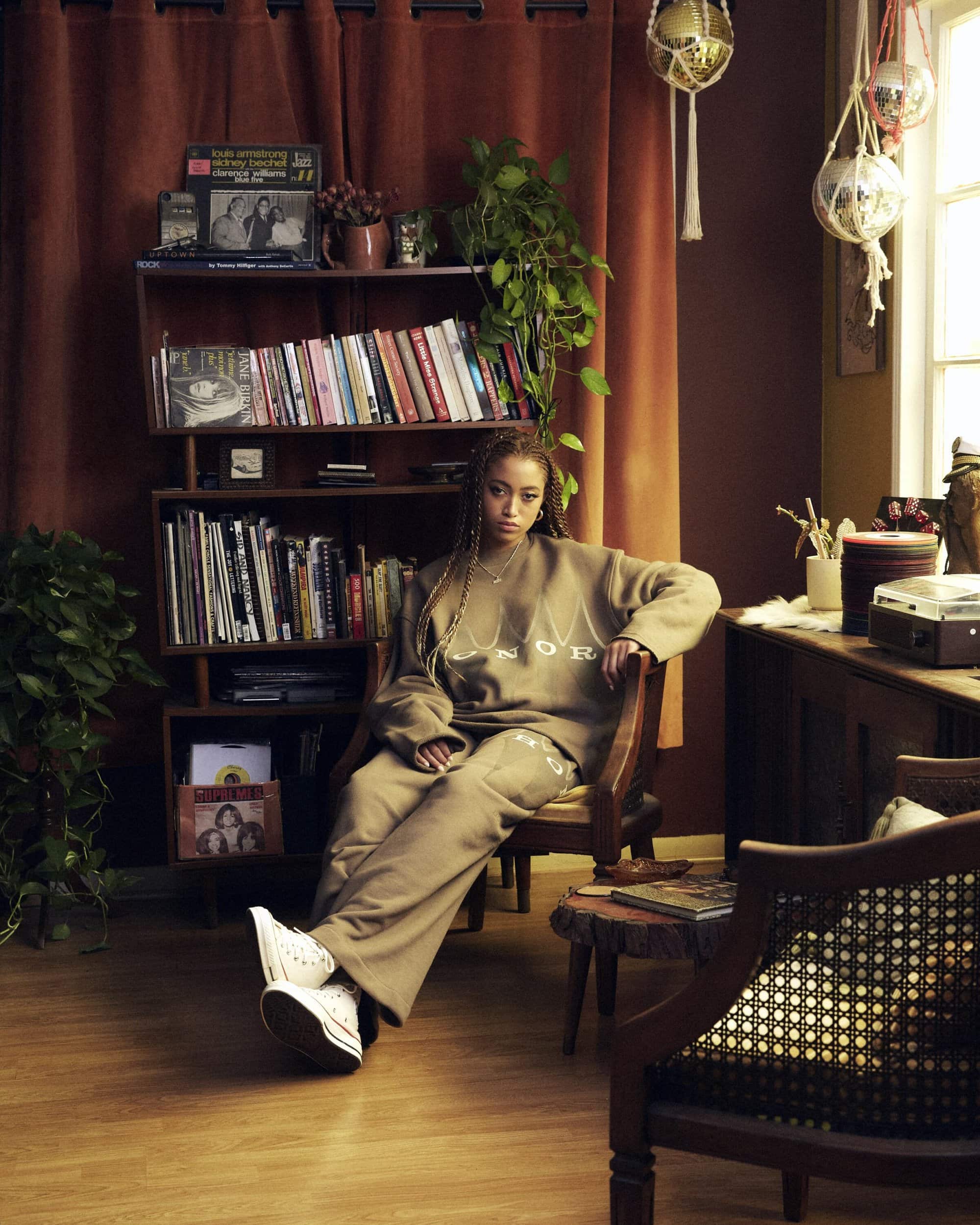 Adult woman in a sweatsuit sits in a vintage-style room with plants, books, and vinyl records in warm, natural light, creating a cozy, lived-in atmosphere.