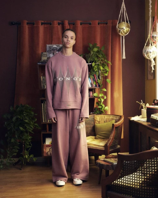 Studio portrait of a young adult standing in casual mauve matching set of a sweatshirt and pants, with bookshelves and home decor.