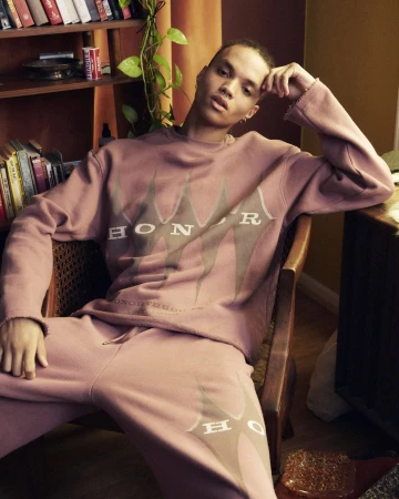 A young adult poses in a mauve sweatsuit with the text 'HONOR' in an indoor setting with a bookshelf and plant.