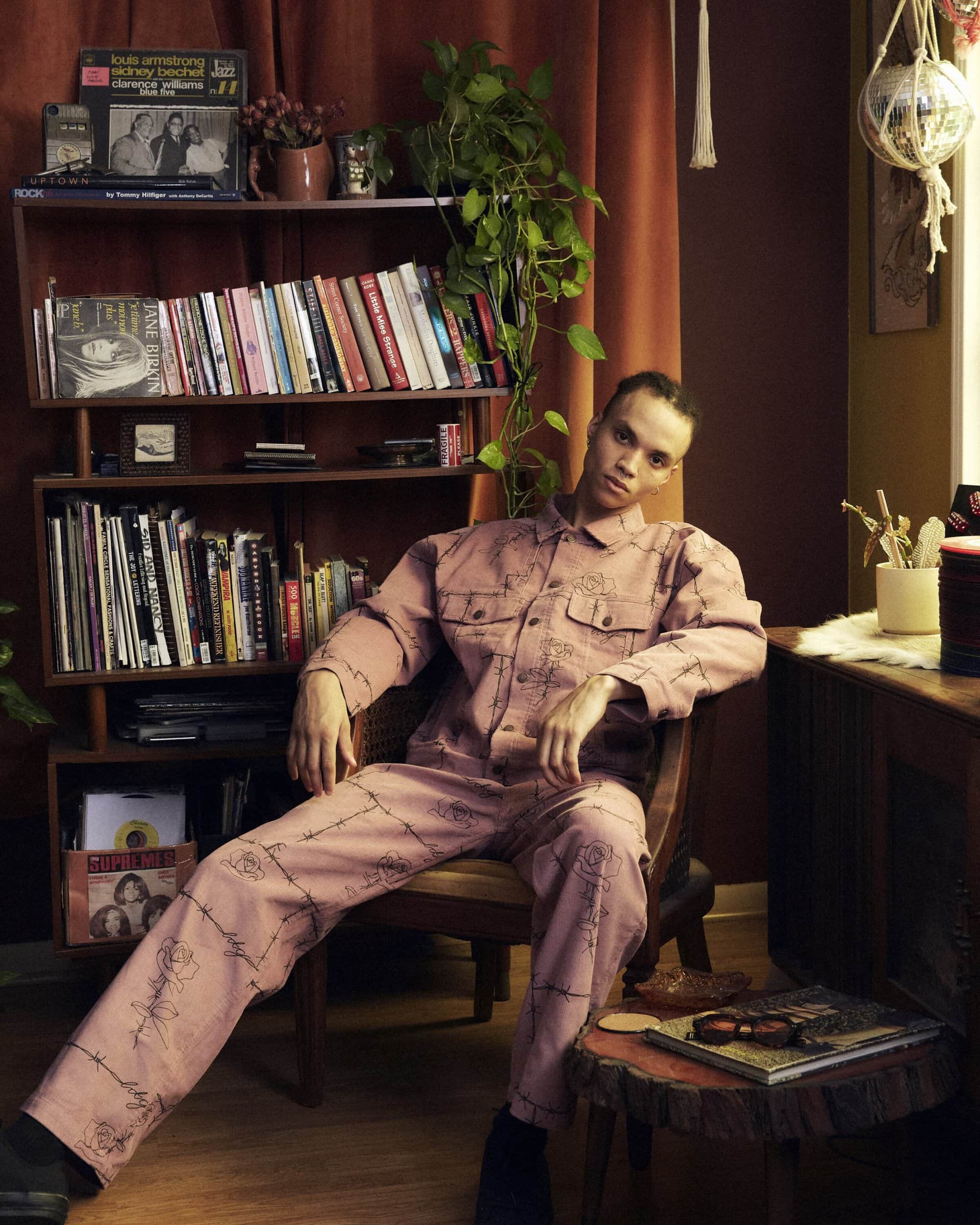 An adult with short dark hair sits on a wooden chair, wearing a pink patterned jumpsuit near a bookshelf in a room with warm lighting.