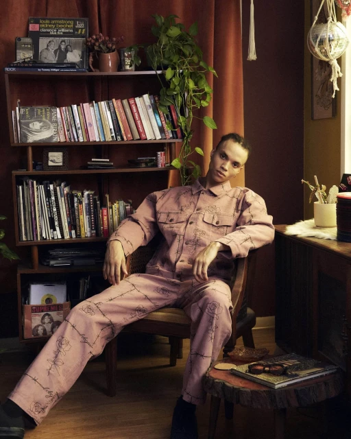 An adult with short dark hair sits on a wooden chair, wearing a pink patterned jumpsuit near a bookshelf in a room with warm lighting.