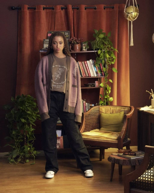 Full-length portrait of an adult woman with braided hair wearing a cardigan and pants, standing in front of a curtain and bookshelf.