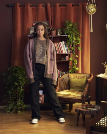 Full-length portrait of an adult woman with braided hair wearing a cardigan and pants, standing in front of a curtain and bookshelf.