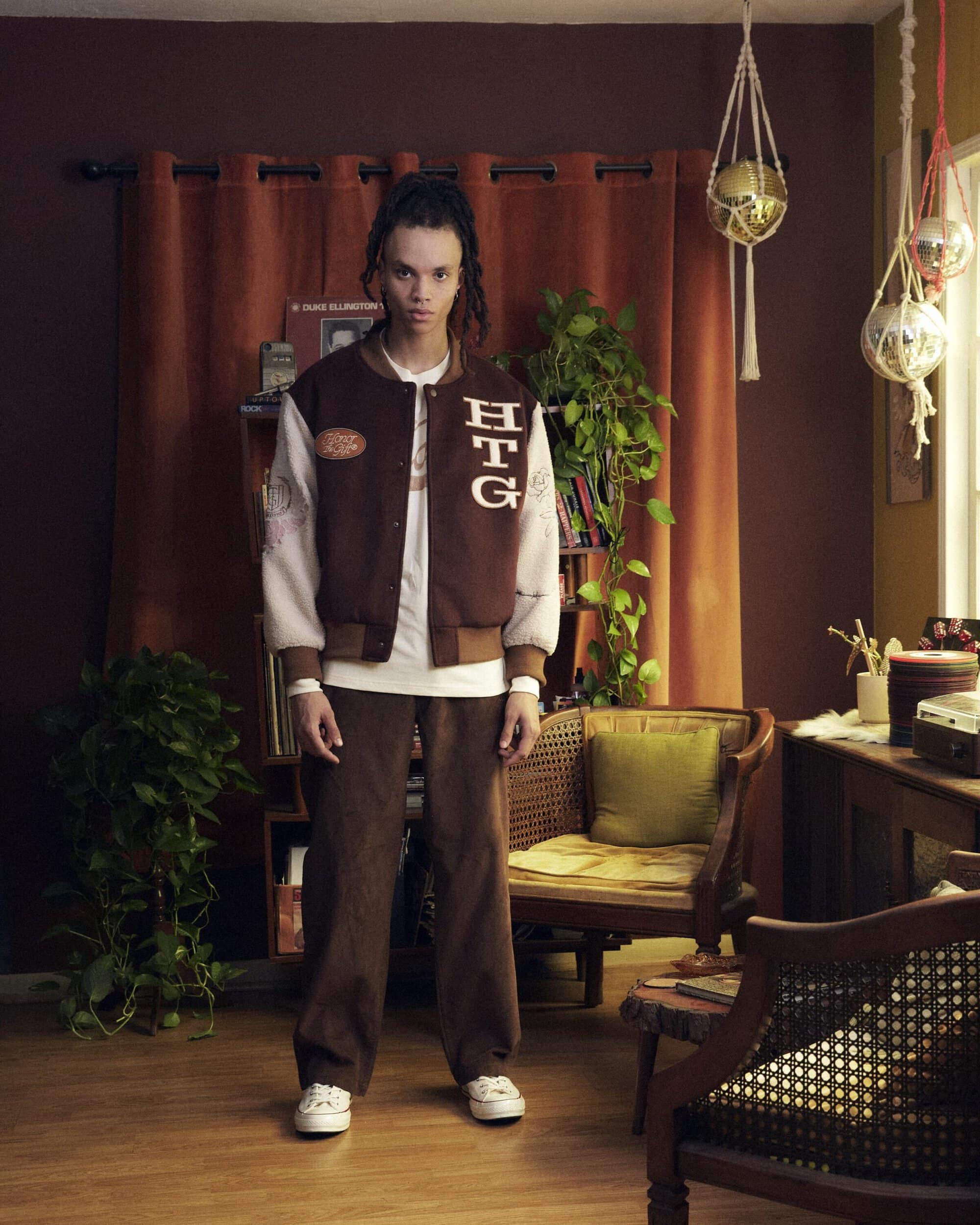 Adult male wearing a brown jacket and pants in an indoor setting with plants and vintage furniture, posing for a full shot.