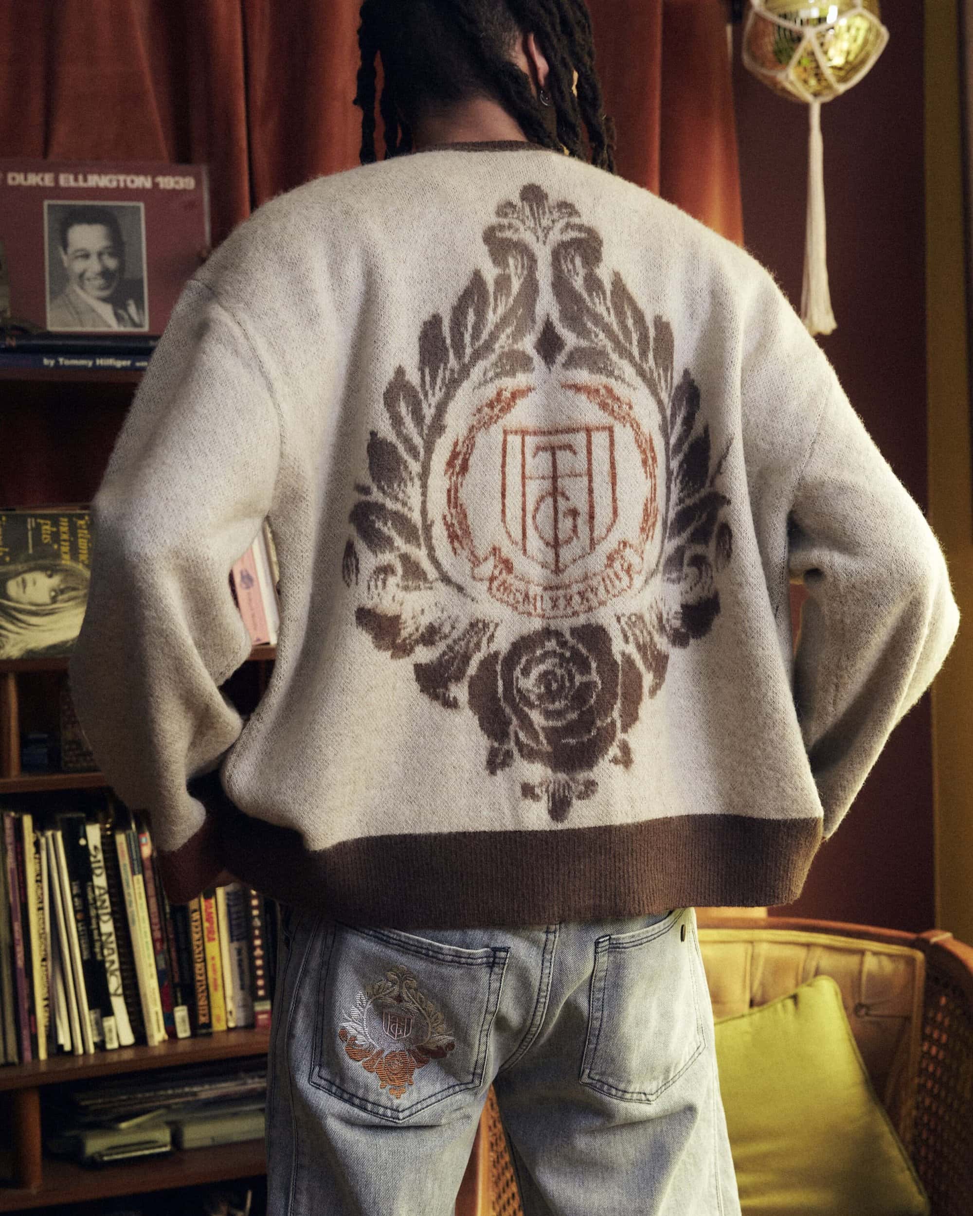 Person wearing a beige knit cardigan with a brown graphic design on the back, light wash jeans with a rose graphic, bookshelf.