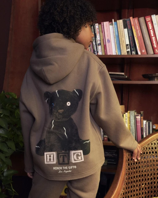 Child poses indoors wearing a brown hooded sweatshirt with a teddy bear graphic, paired with matching sweatpants and a bookshelf backdrop.