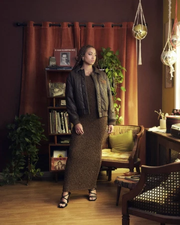 A woman stands in a living room wearing a brown textured maxi dress and a quilted dark bomber jacket, looking away from the camera.