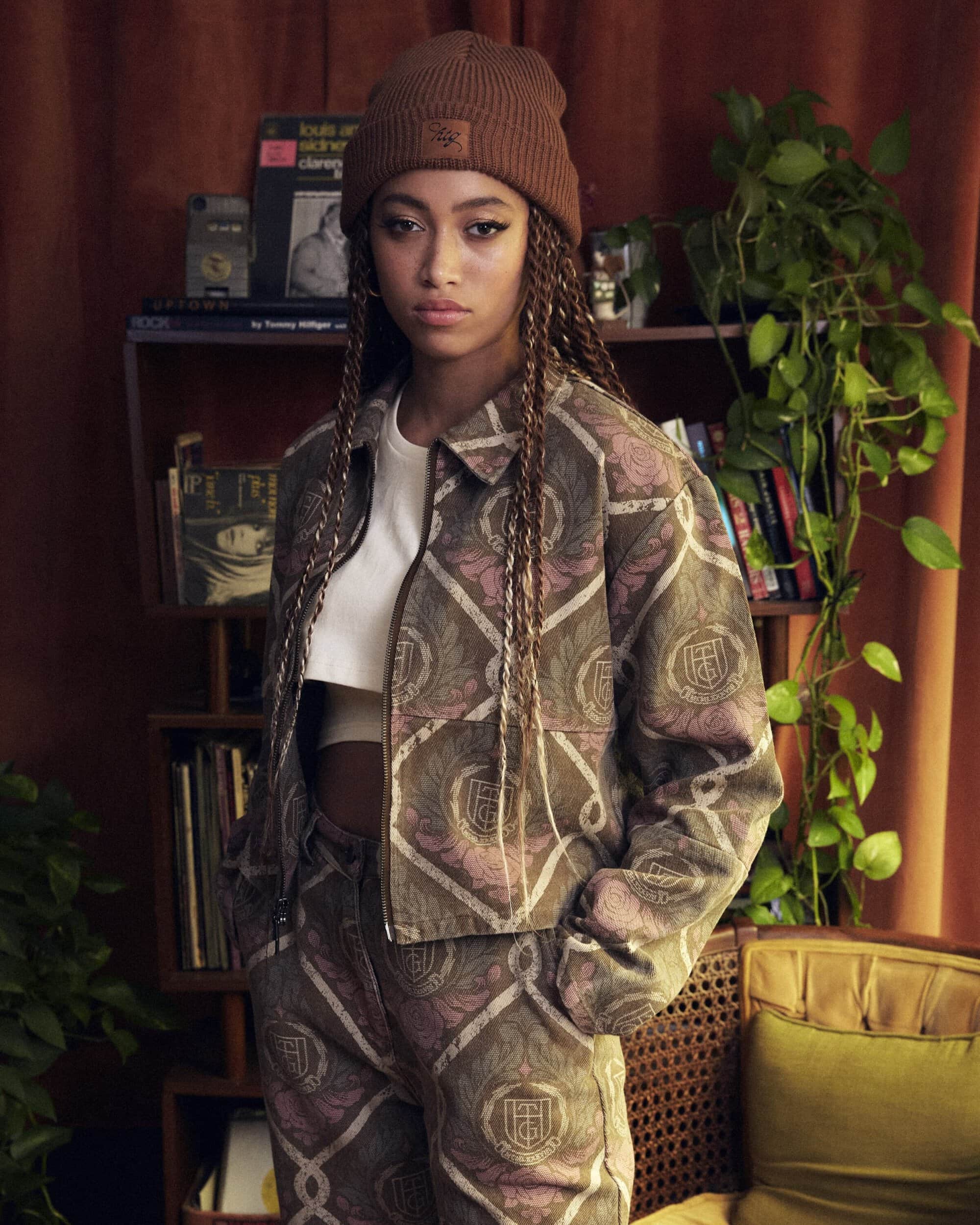 Young woman in a patterned jacket and matching pants poses indoors in front of a shelf holding records and plants.