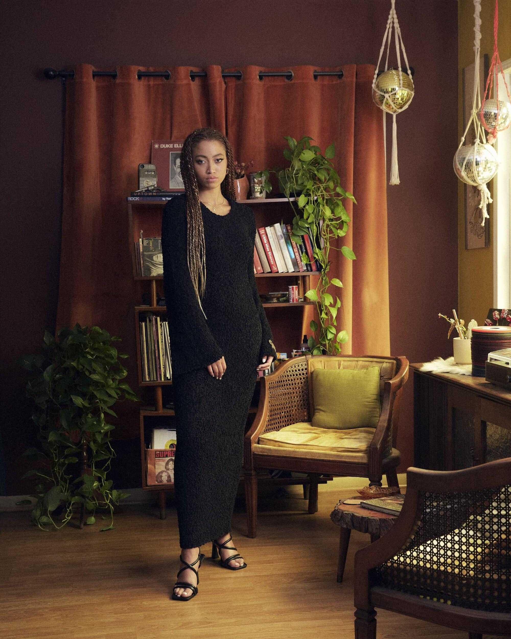 Woman in a black knit dress stands in a living room with houseplants, records, and rattan chairs, creating a cozy, vintage-inspired setting.