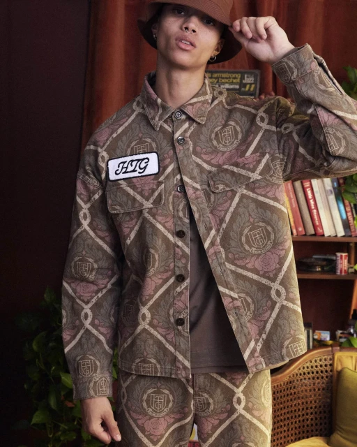 Young adult in a matching brown patterned jacket and pants, posing indoors in front of a bookshelf. Wearing a matching brown bucket hat.