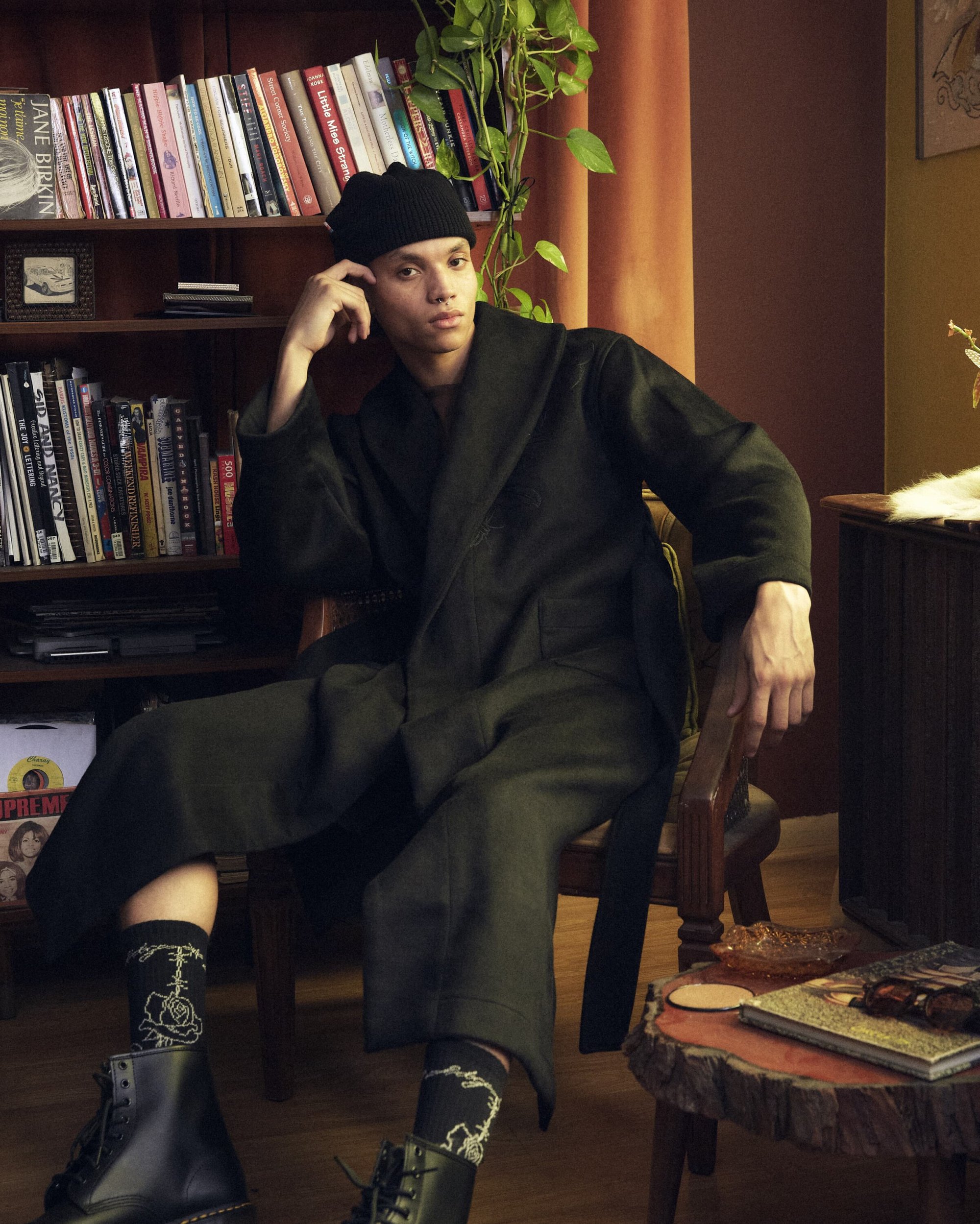 Model seated in a wooden chair wearing a dark robe coat, black beanie, graphic socks, and boots beside a bookshelf.