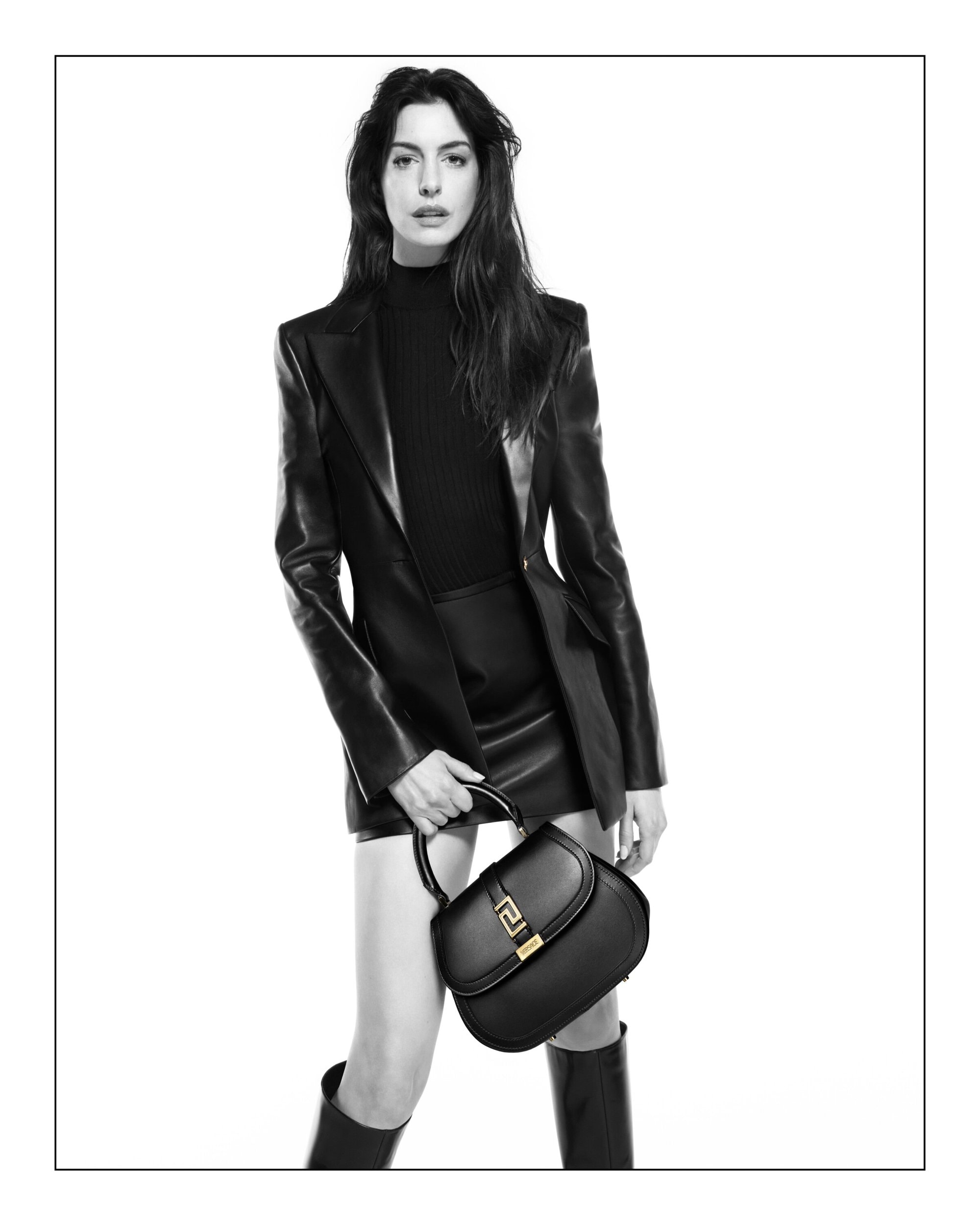 Anne Hathaway poses in a black leather jacket and matching mini skirt, accessorized with a black Versace handbag in a studio setting.