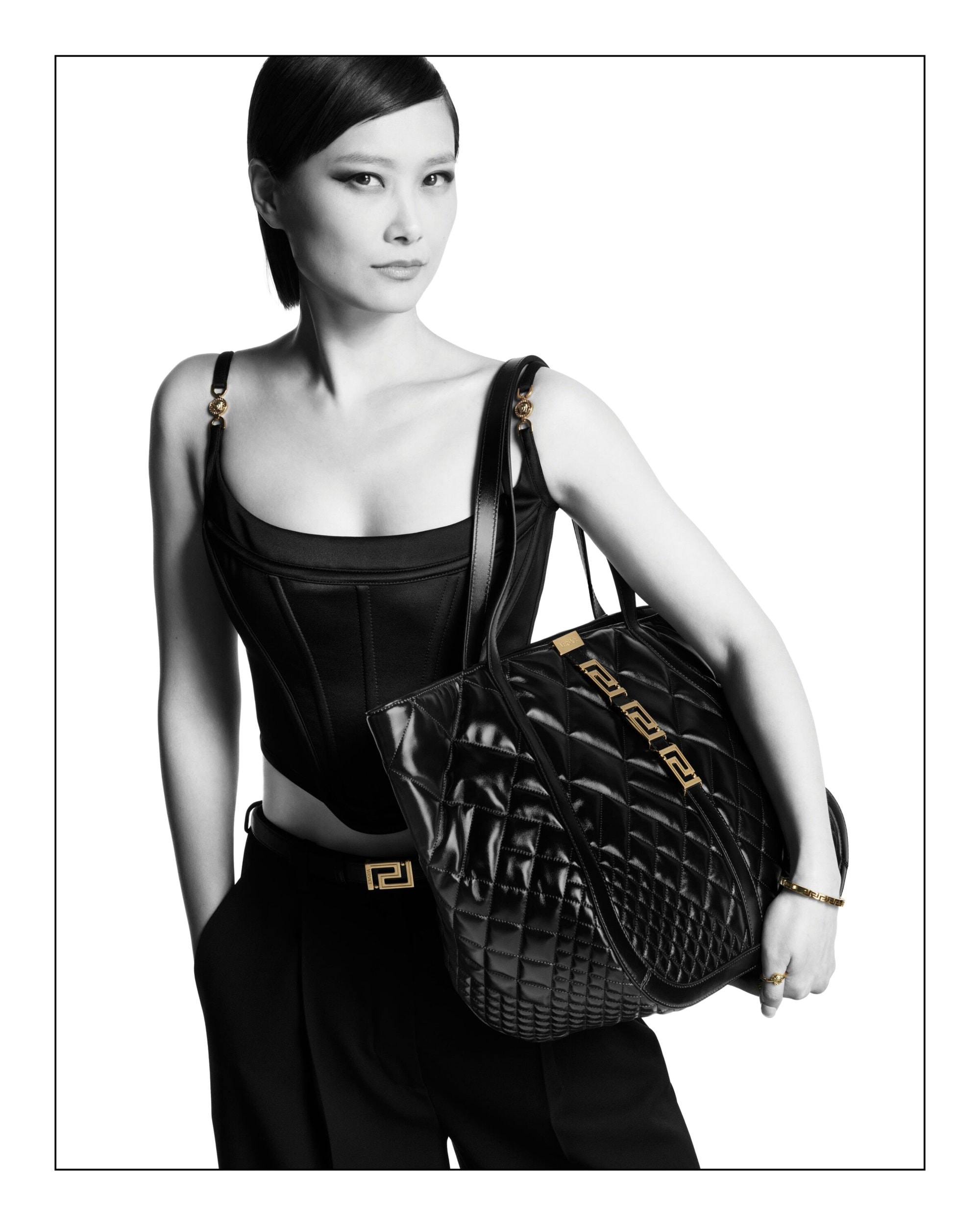 Asian woman in black corset and trousers carries a quilted leather Versace tote bag in a studio product shot on white background.