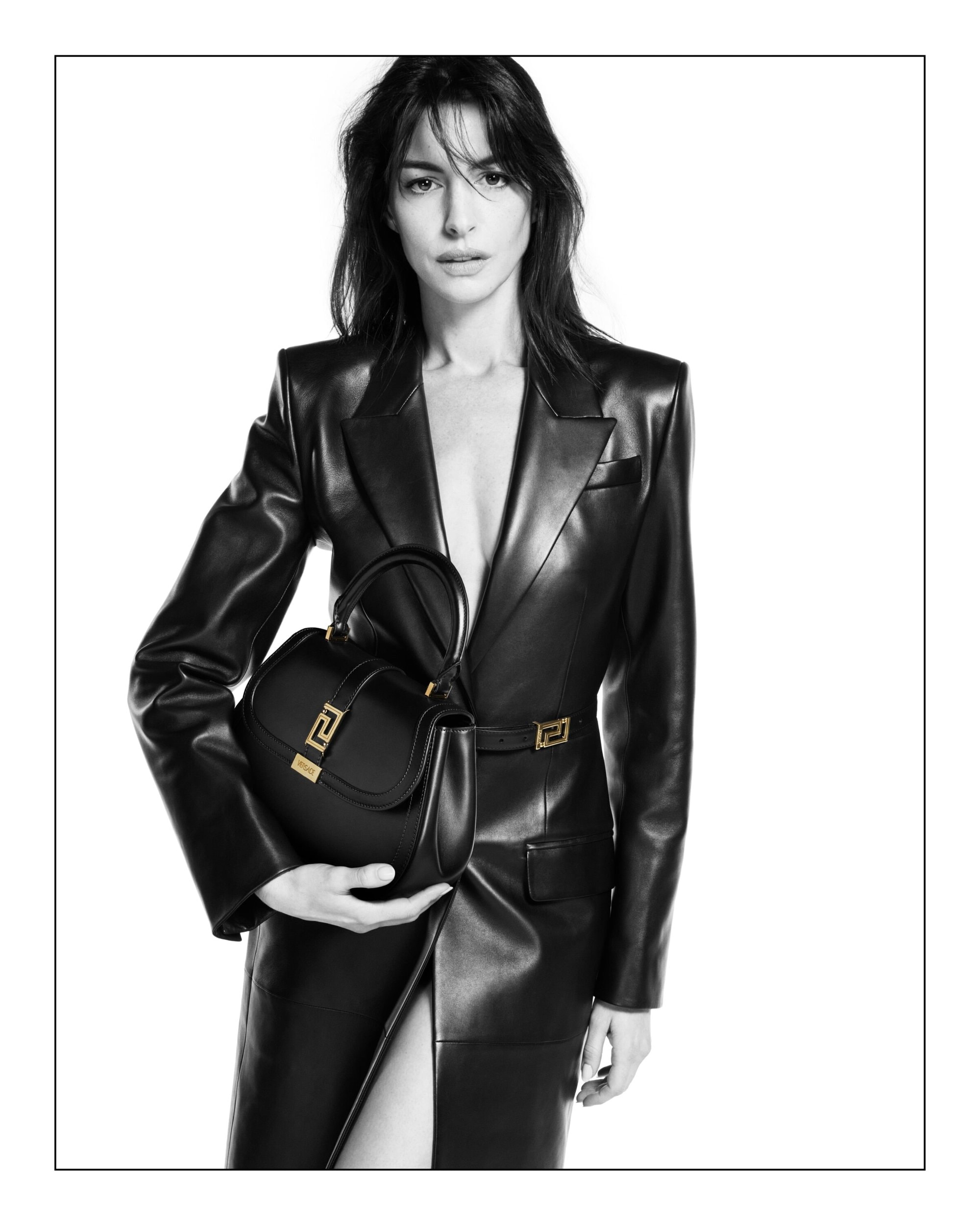 A woman in a black leather dress holds a black leather handbag with gold accents, against a bright white studio background.