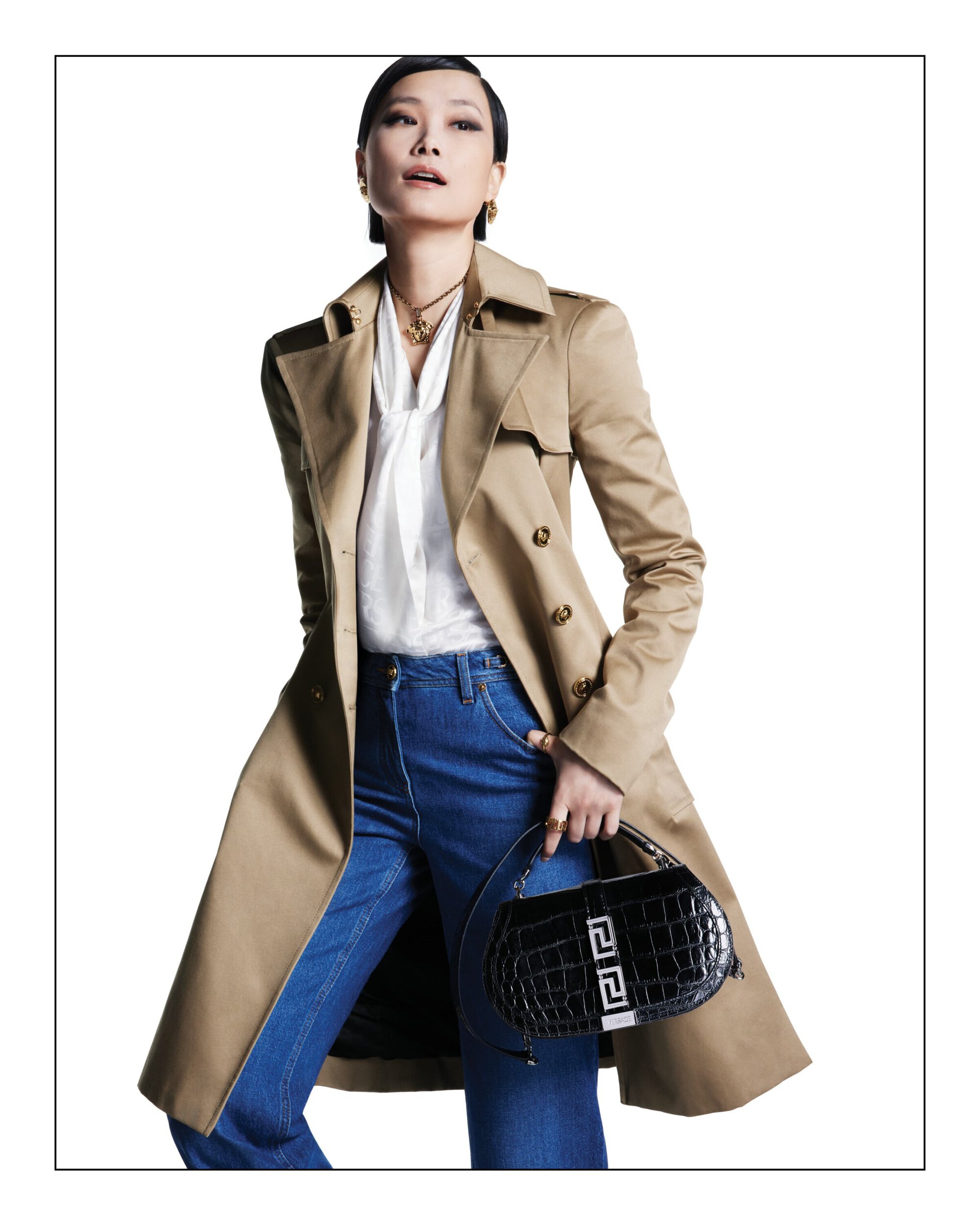 Elegant woman in a trench coat, jeans, and handbag posing against a white backdrop in a fashion editorial studio shot.