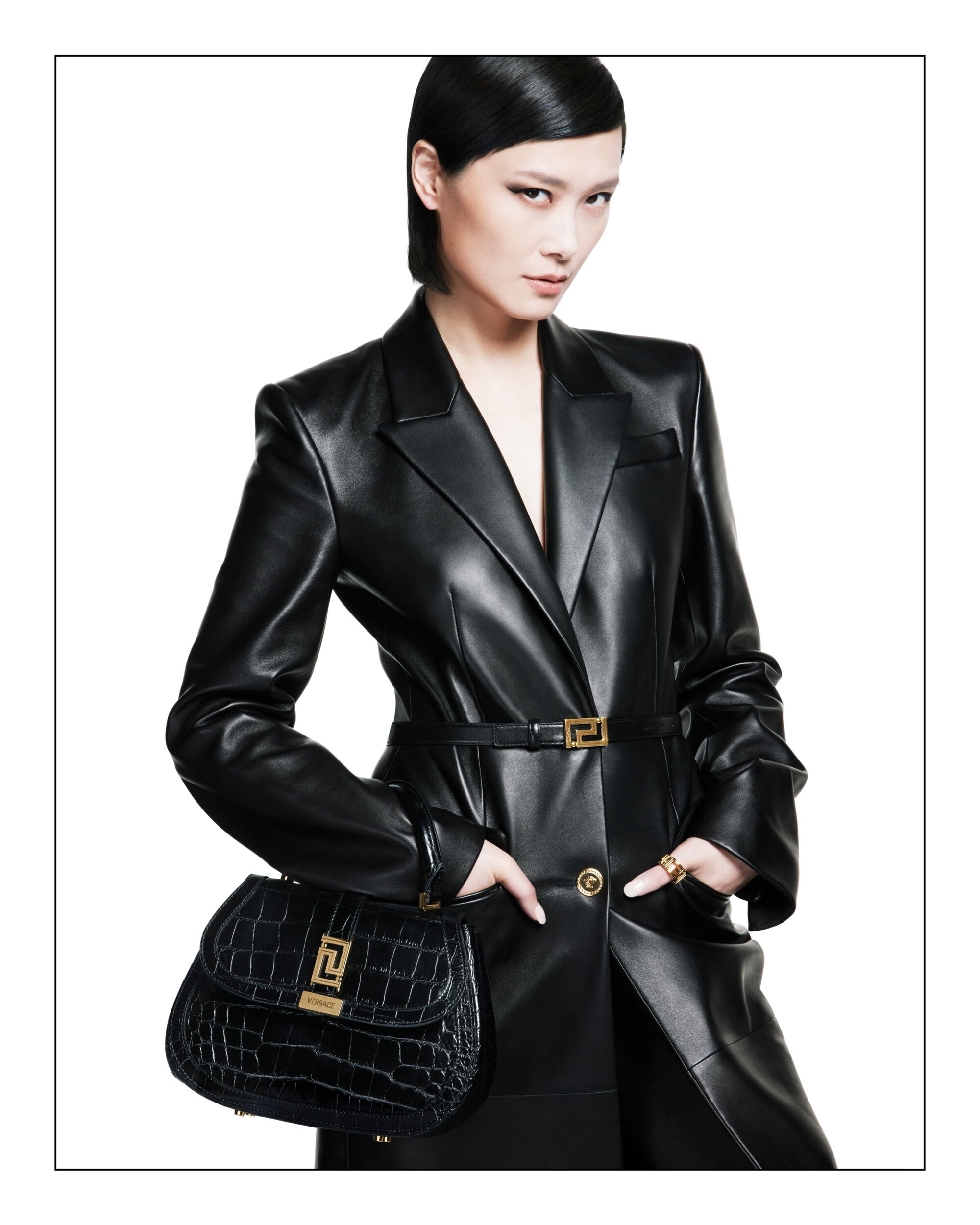 Fashion shot of an adult female in a black leather coat dress with a matching purse against a white background in studio lighting.