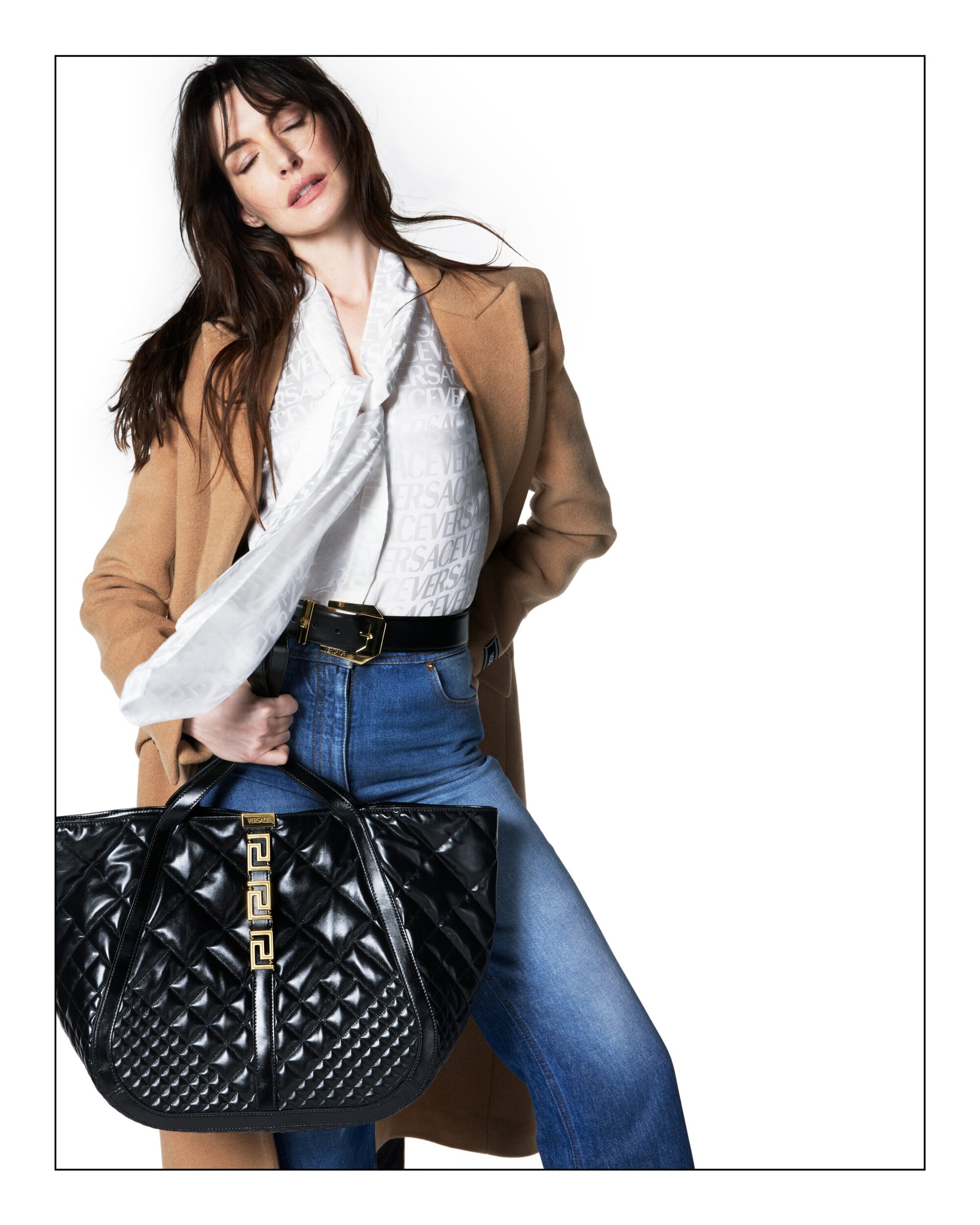 Brunette woman posing with a black quilted Versace handbag in an off-center studio shot. She's wearing jeans, a blouse, and a coat.