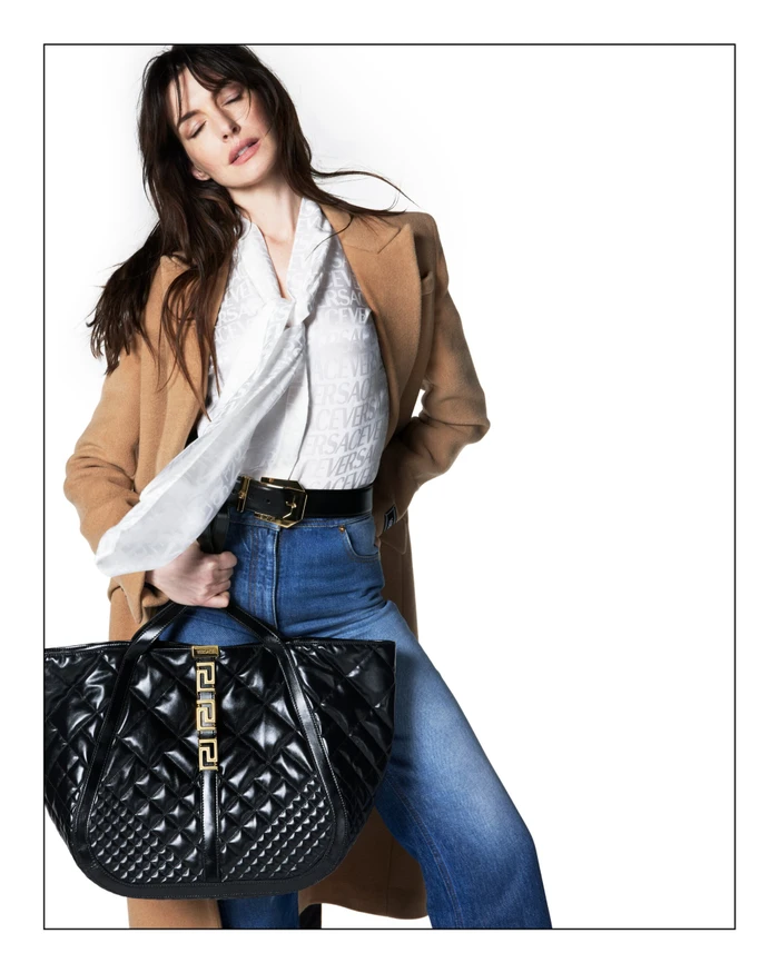 Anne Hathaway wearing a brown coat and blue jeans holding a black quilted handbag against a plain white background.