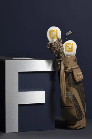 Elevated studio shot showcases a tan Fendi golf bag, golf clubs, and head covers next to a silver Fendi F on a gray surface against a blue backdrop.