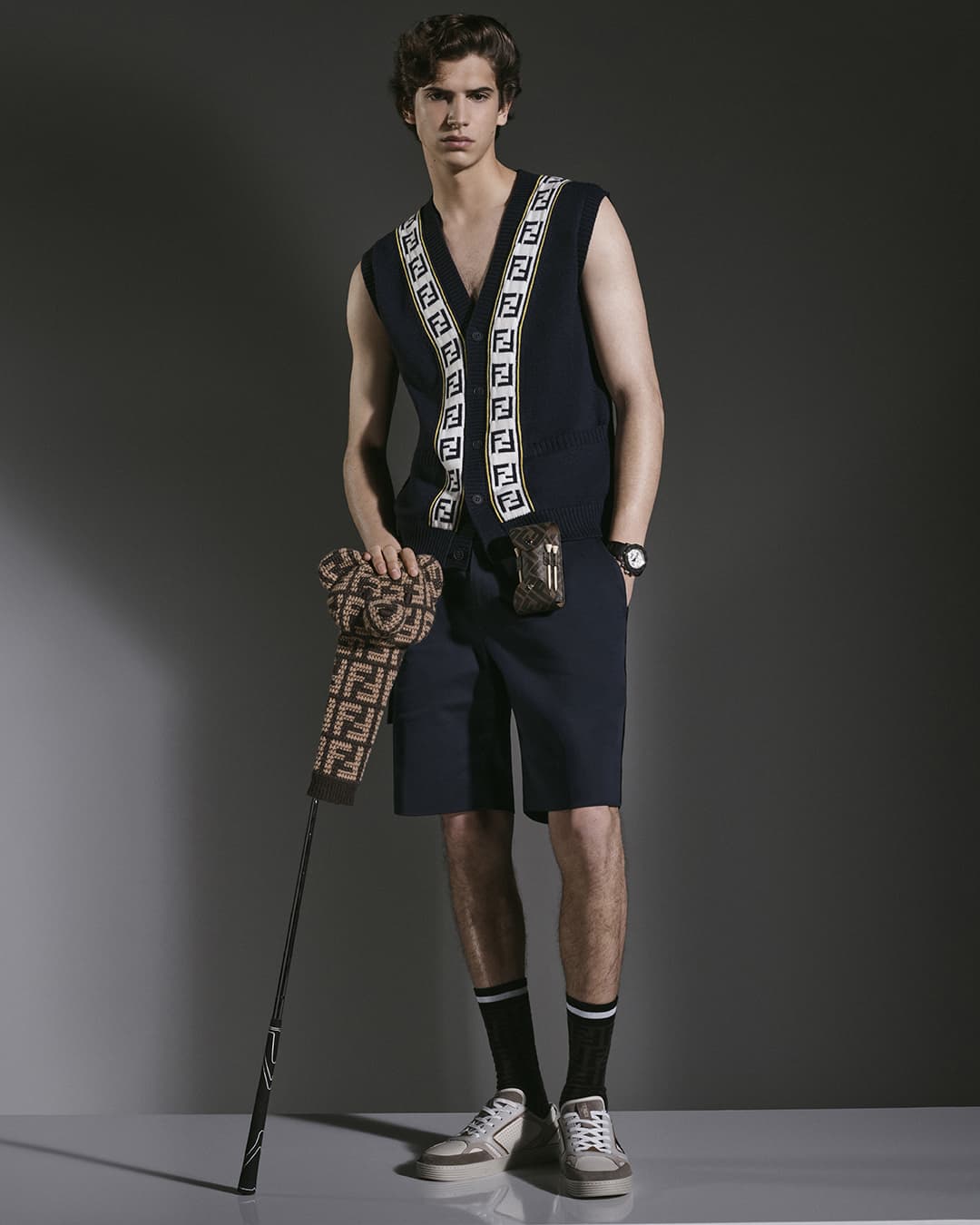 A young man poses indoors wearing a navy vest, shorts, and sneakers, holding a golf club with a patterned golf-club cover.