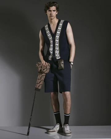 A young man poses indoors wearing a navy vest, shorts, and sneakers, holding a golf club with a patterned golf-club cover.