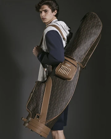 Male model carrying a brown monogram leather golf bag over his shoulder against a dark studio background.