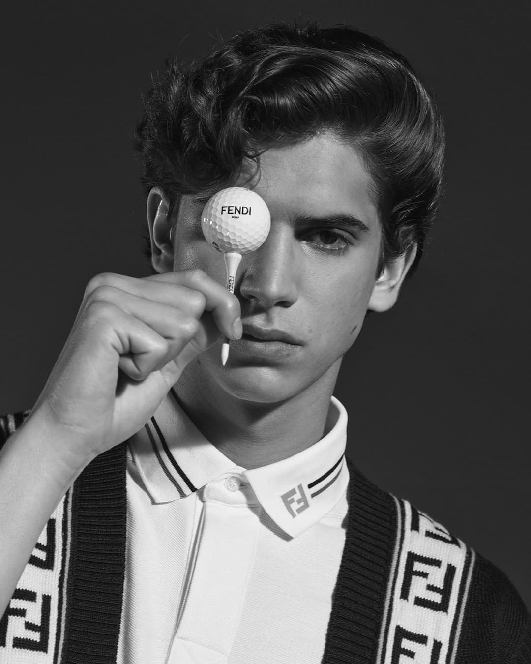 Monochrome portrait of a young adult male holding a golf ball with branding over one eye in a photo studio setting.