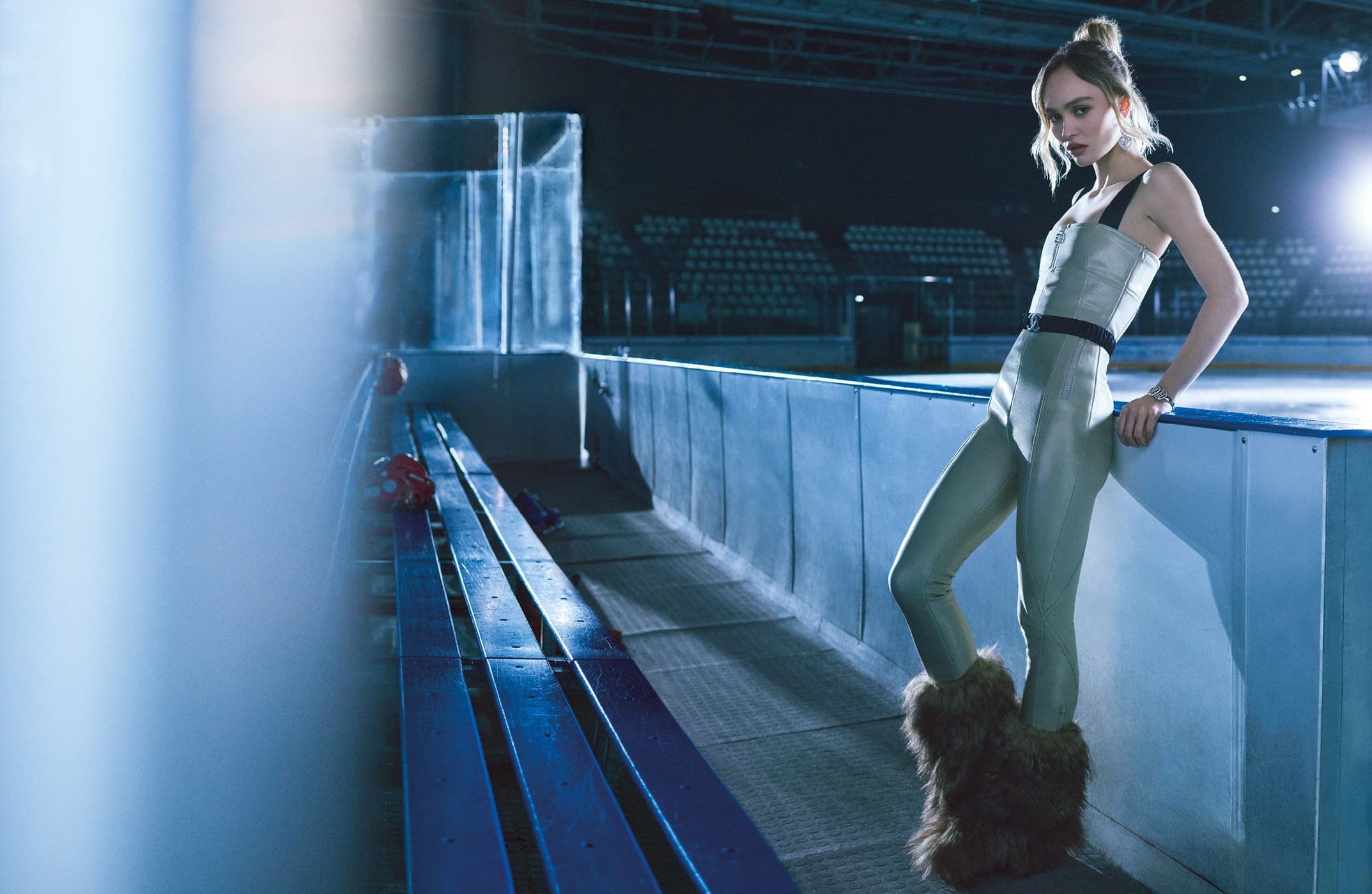 A woman in a pale green jumpsuit and fur boots leans against an ice rink barrier with stadium seating in the background.