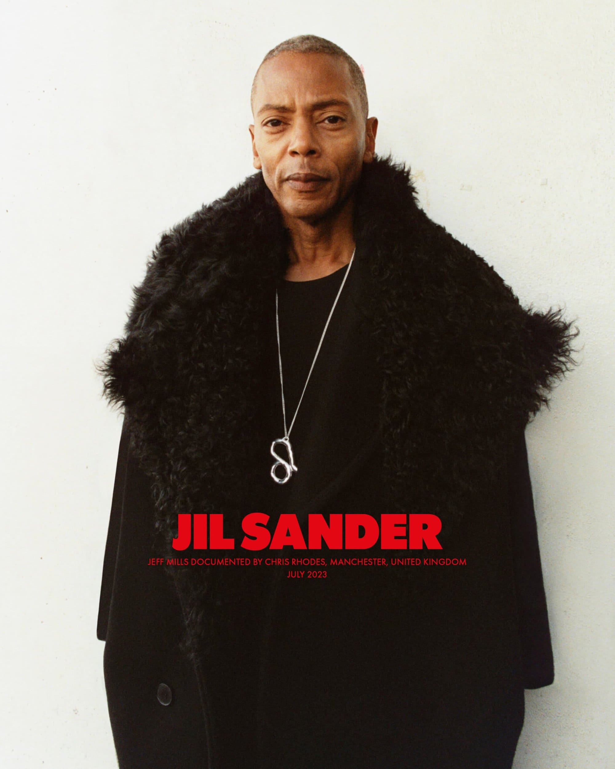 A portrait shot captures a distinguished man wearing a black coat with fur collar and silver necklace, against a plain white background.