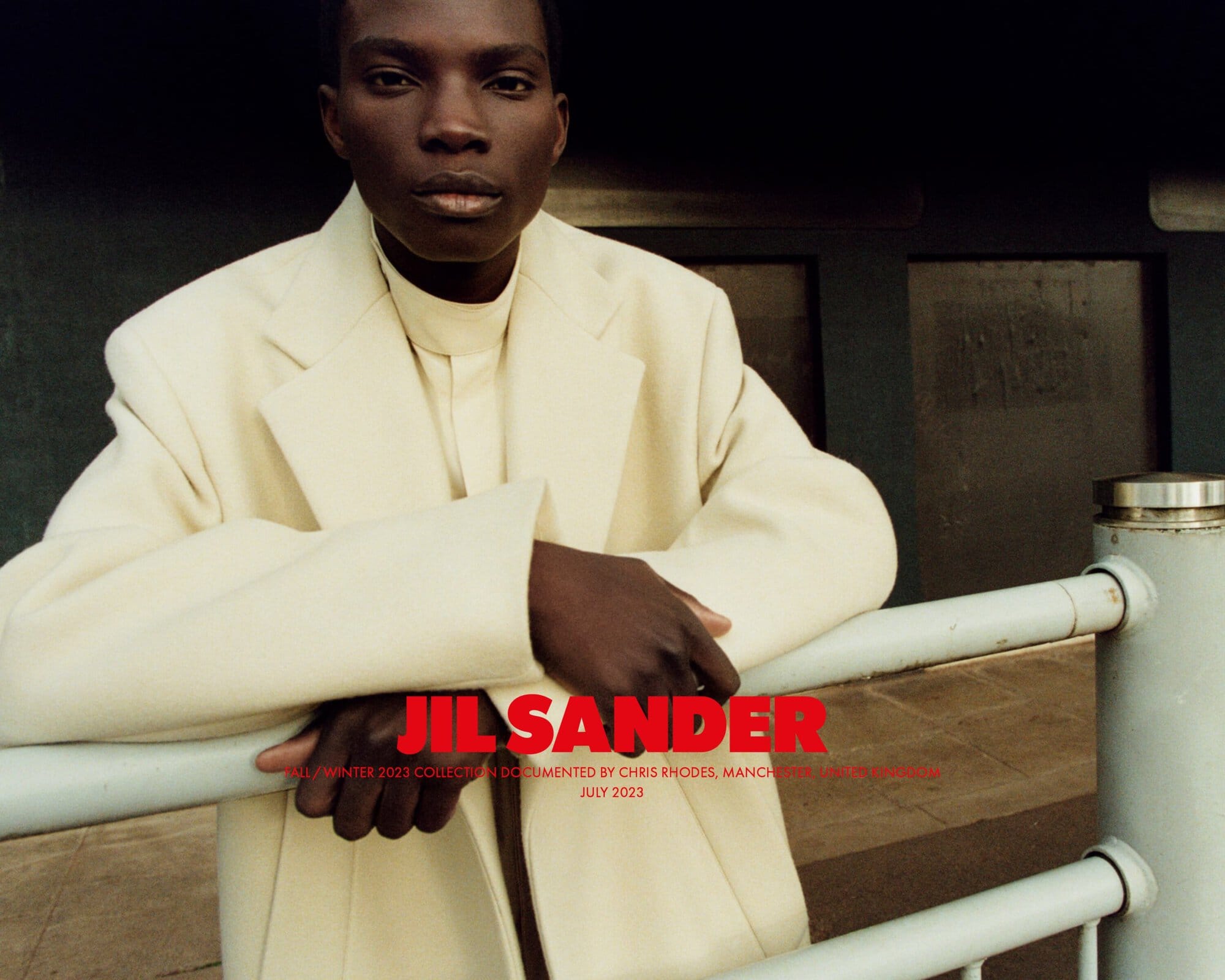 An adult male model of African descent poses in a cream-colored jacket and shirt next to a metal railing in front of a dark background.