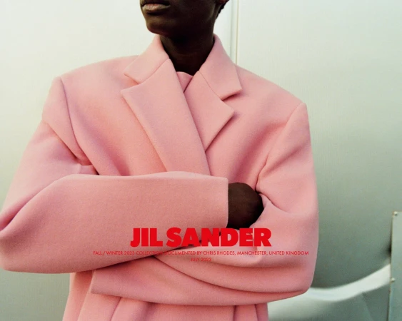 Dark-skinned model wears a pink Jil Sander Fall/Winter 2023 overcoat with folded arms, posing indoors against a gray background with text overlay.