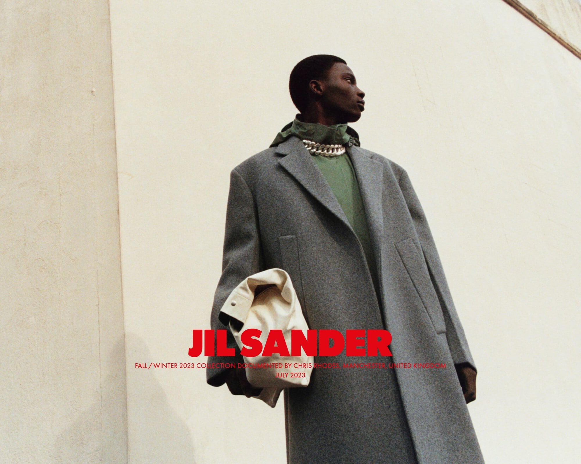 An African American male model showcases Jil Sander Fall/Winter 2023 collection, wearing a grey coat, green top, and chain necklace, outdoors.