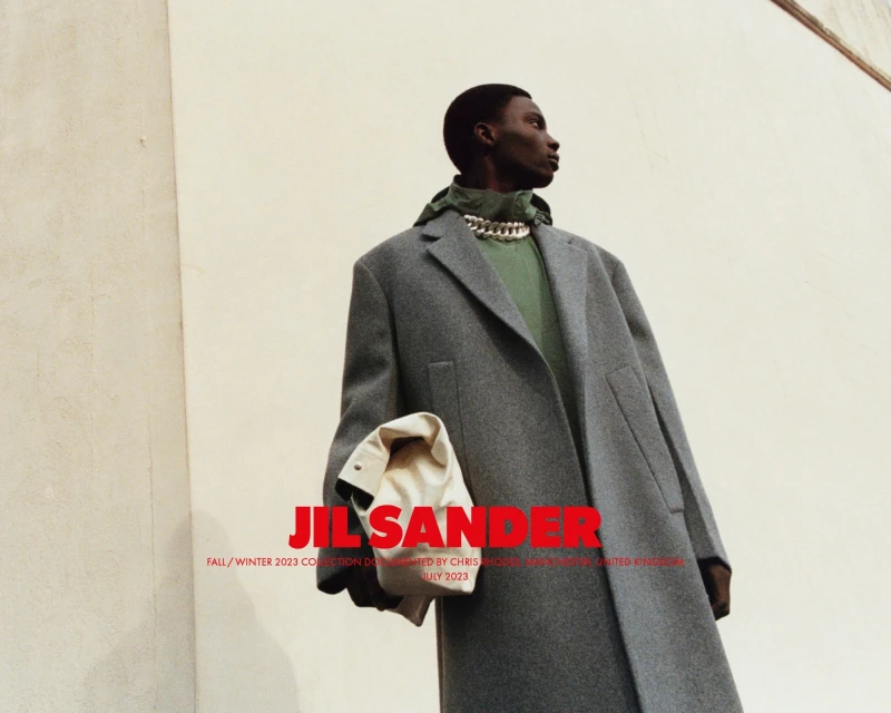 An African American male model showcases Jil Sander Fall/Winter 2023 collection, wearing a grey coat, green top, and chain necklace, outdoors.