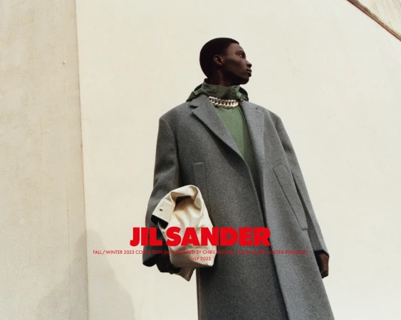 An African American male model showcases Jil Sander Fall/Winter 2023 collection, wearing a grey coat, green top, and chain necklace, outdoors.