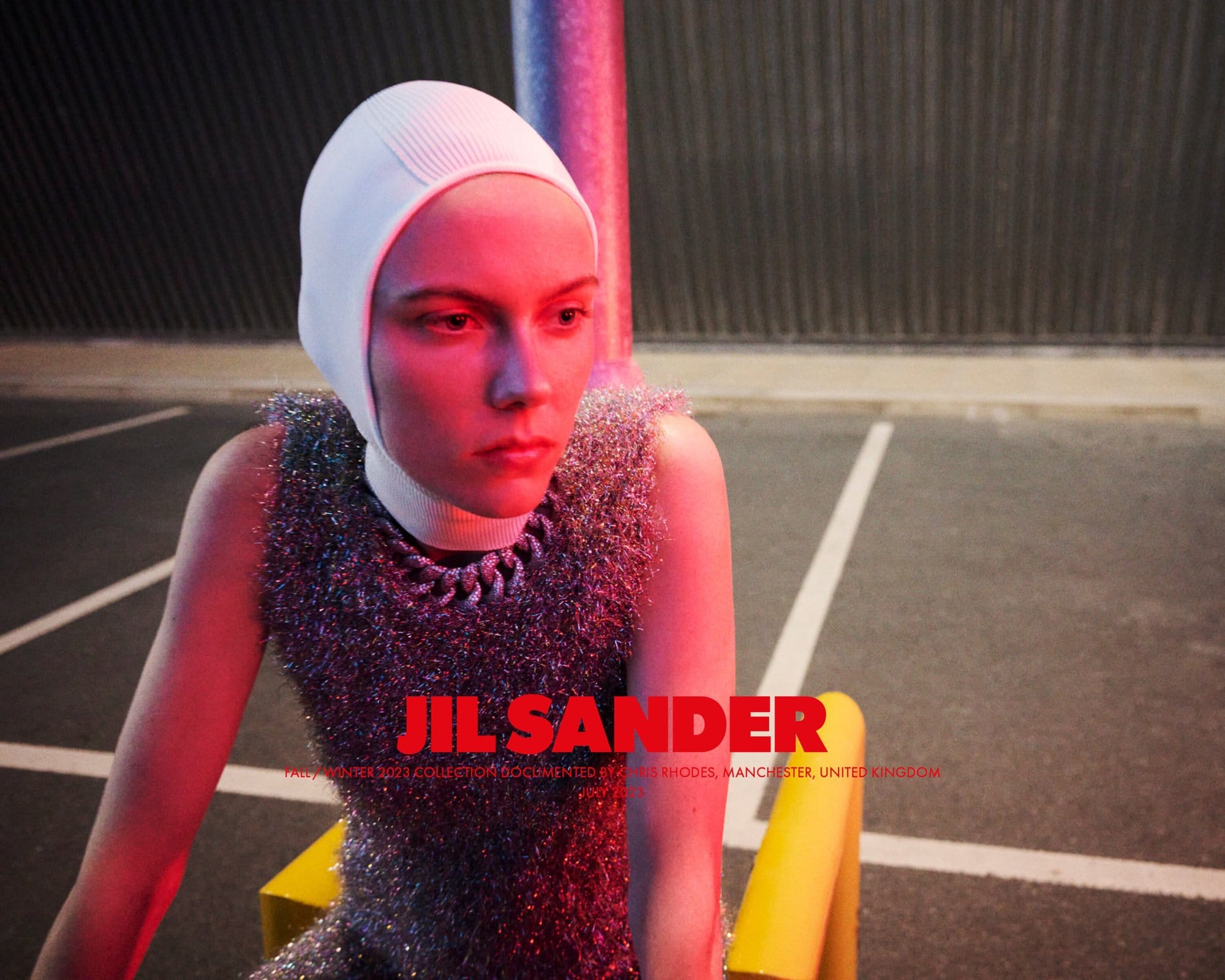 A woman in a futuristic white head covering and a silver, shimmery top sitting on a yellow structure with a concrete wall background.